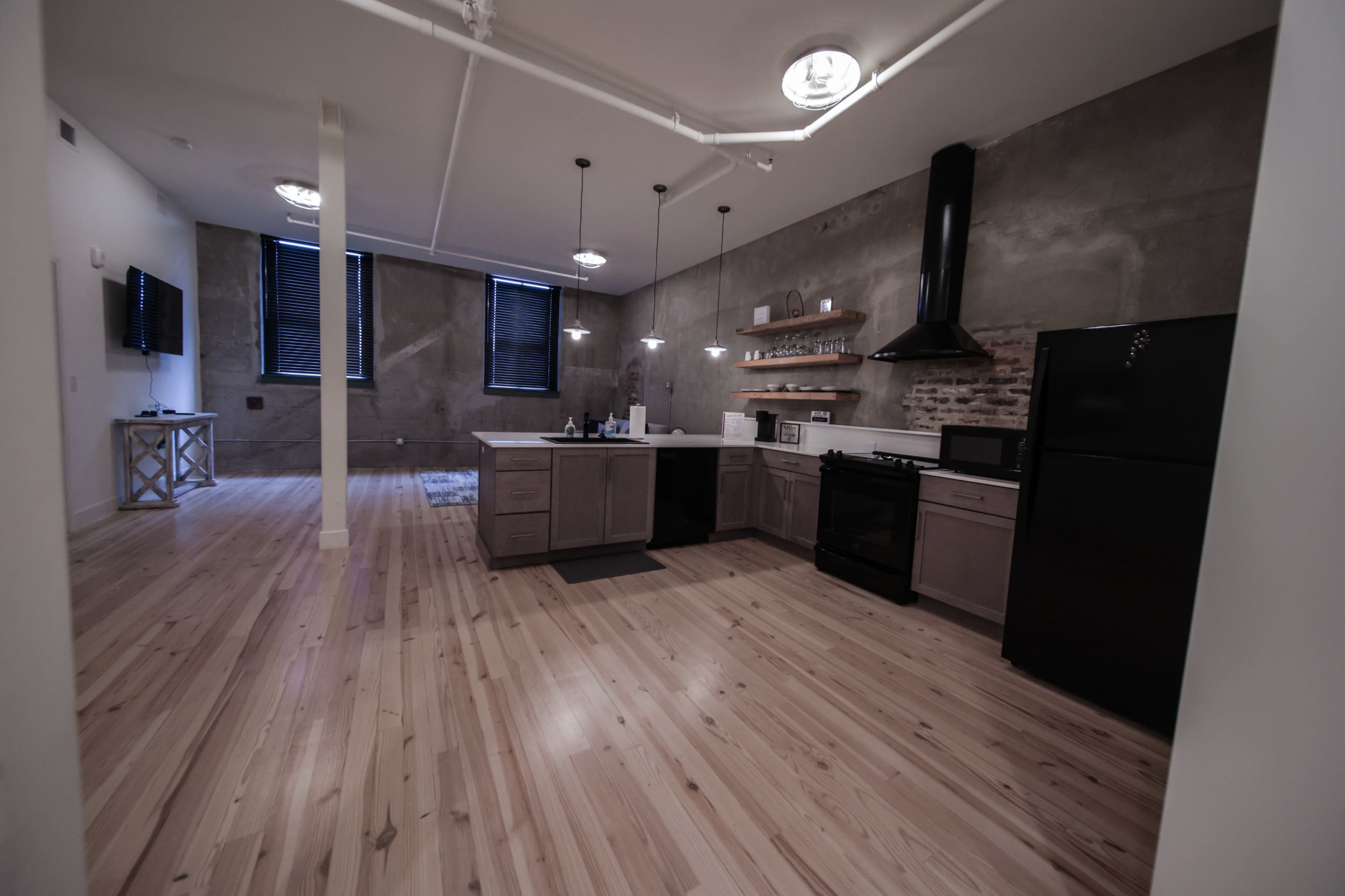 The image shows a spacious kitchen area with modern appliances, a black stove, and light wooden flooring in an industrial-style room with exposed concrete walls.