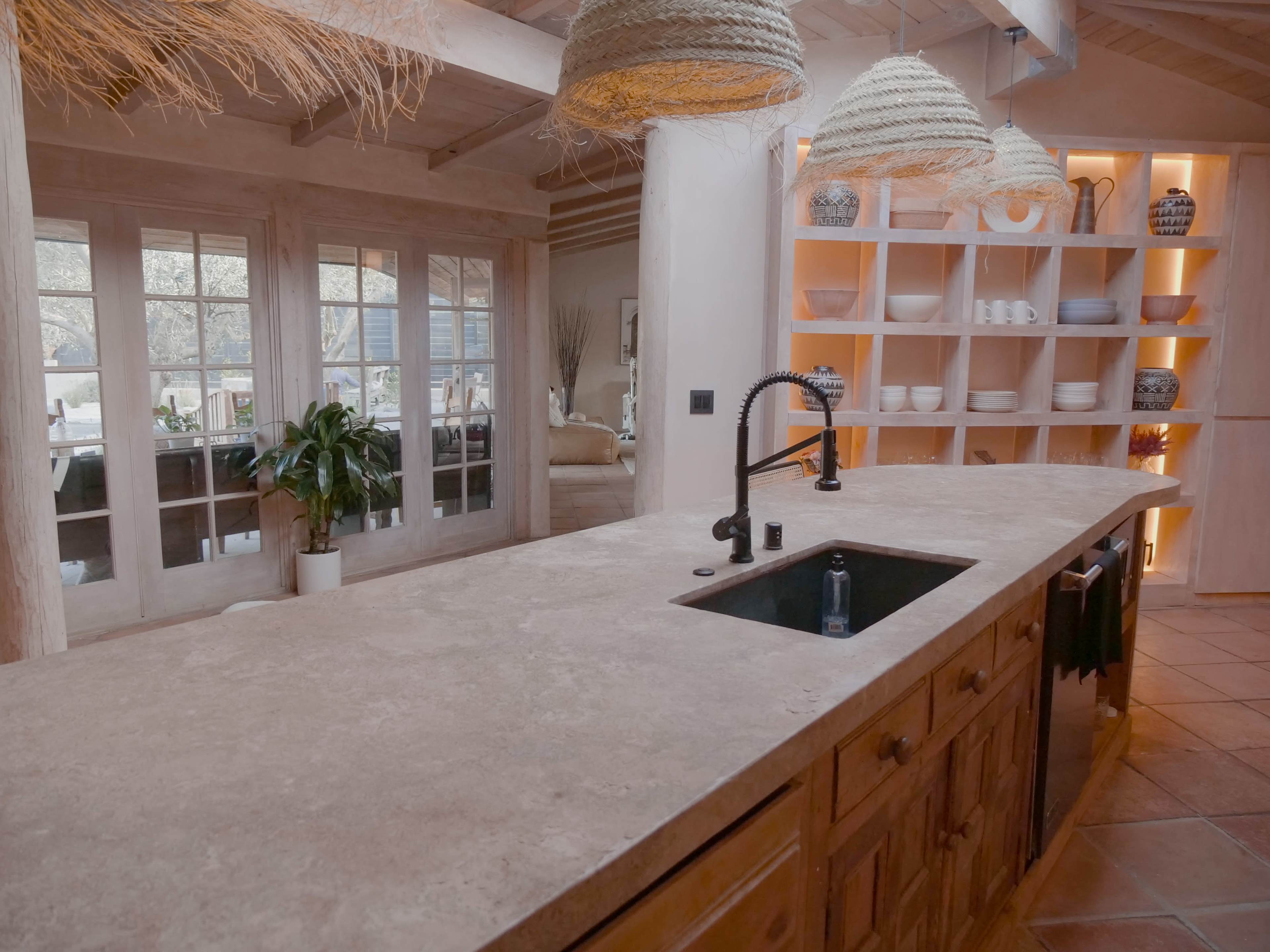 A modern kitchen features a stone countertop with a black sink and decorative pendant lights above, while shelves display white dishware and a view of a living area is visible through large windows.