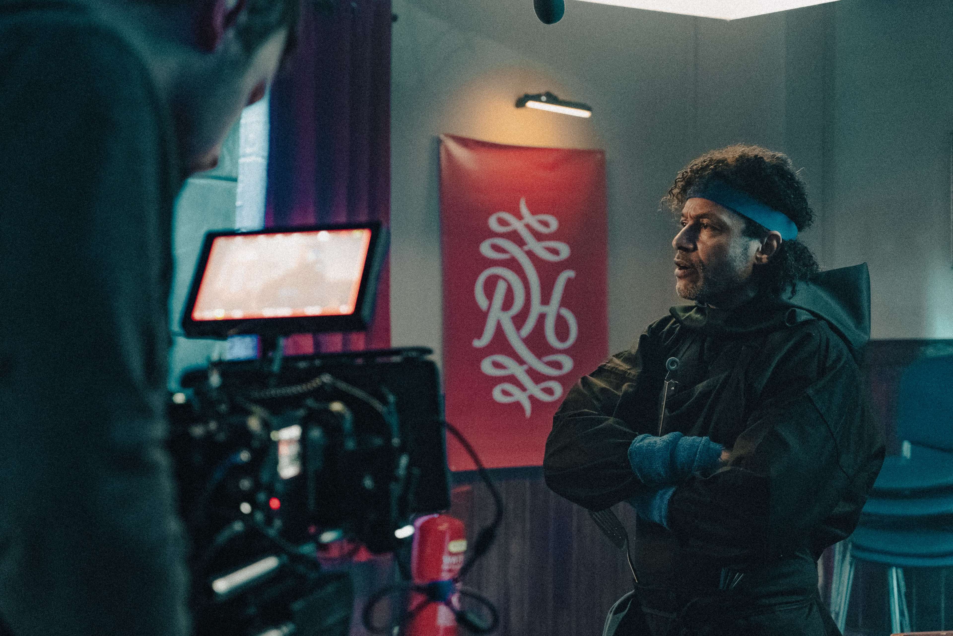A man with curly hair, wearing a blue headband and a black outfit, stands with his arms crossed in front of a camera setup and a decorative banner on the wall.