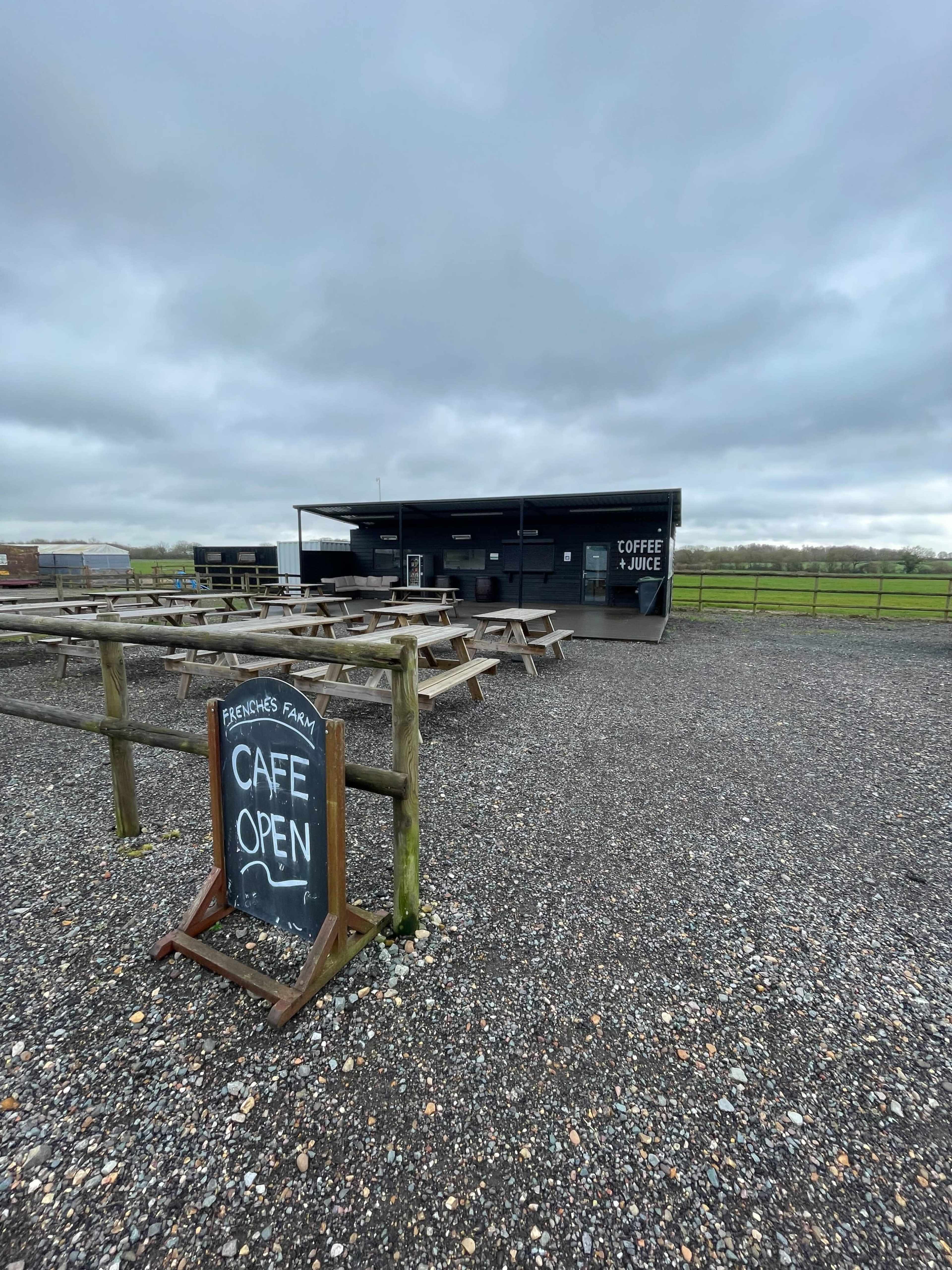 The image shows an outdoor cafe with several picnic tables and a sign indicating that the cafe is open, set against a cloudy sky.