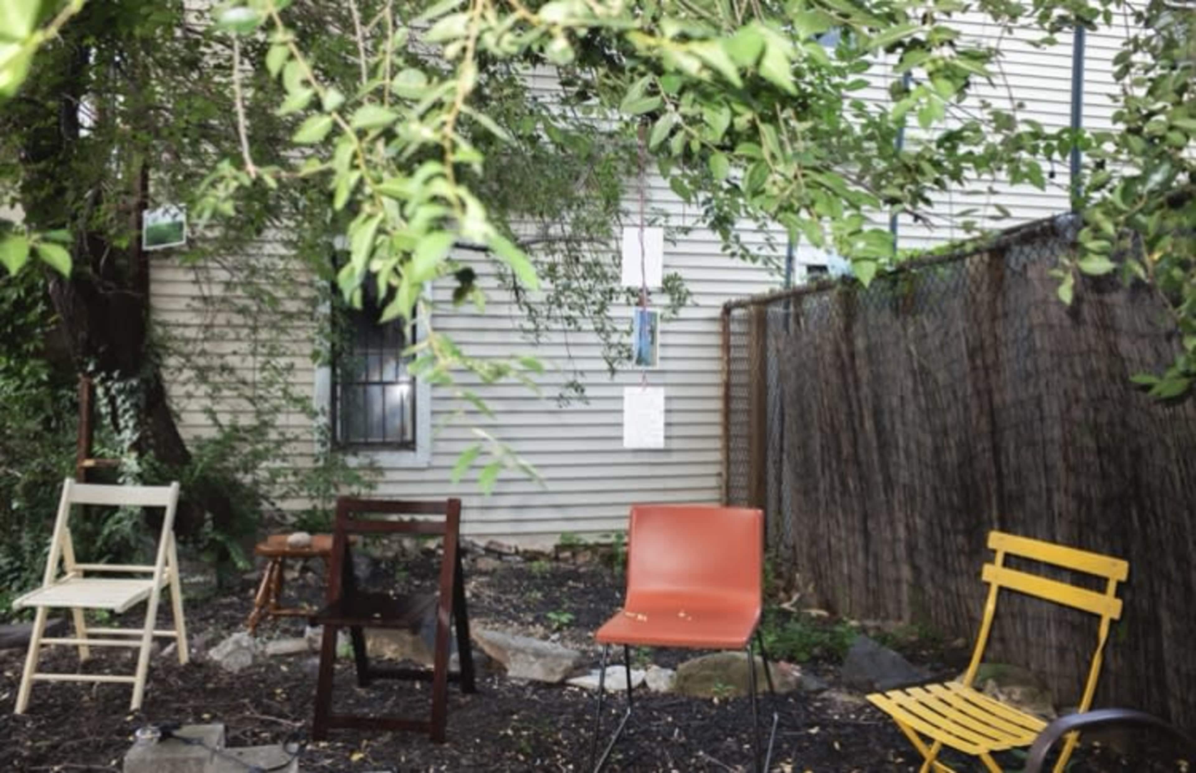 A small outdoor space features a collection of various chairs arranged among rocks and greenery beside a wooden fence and a house.
