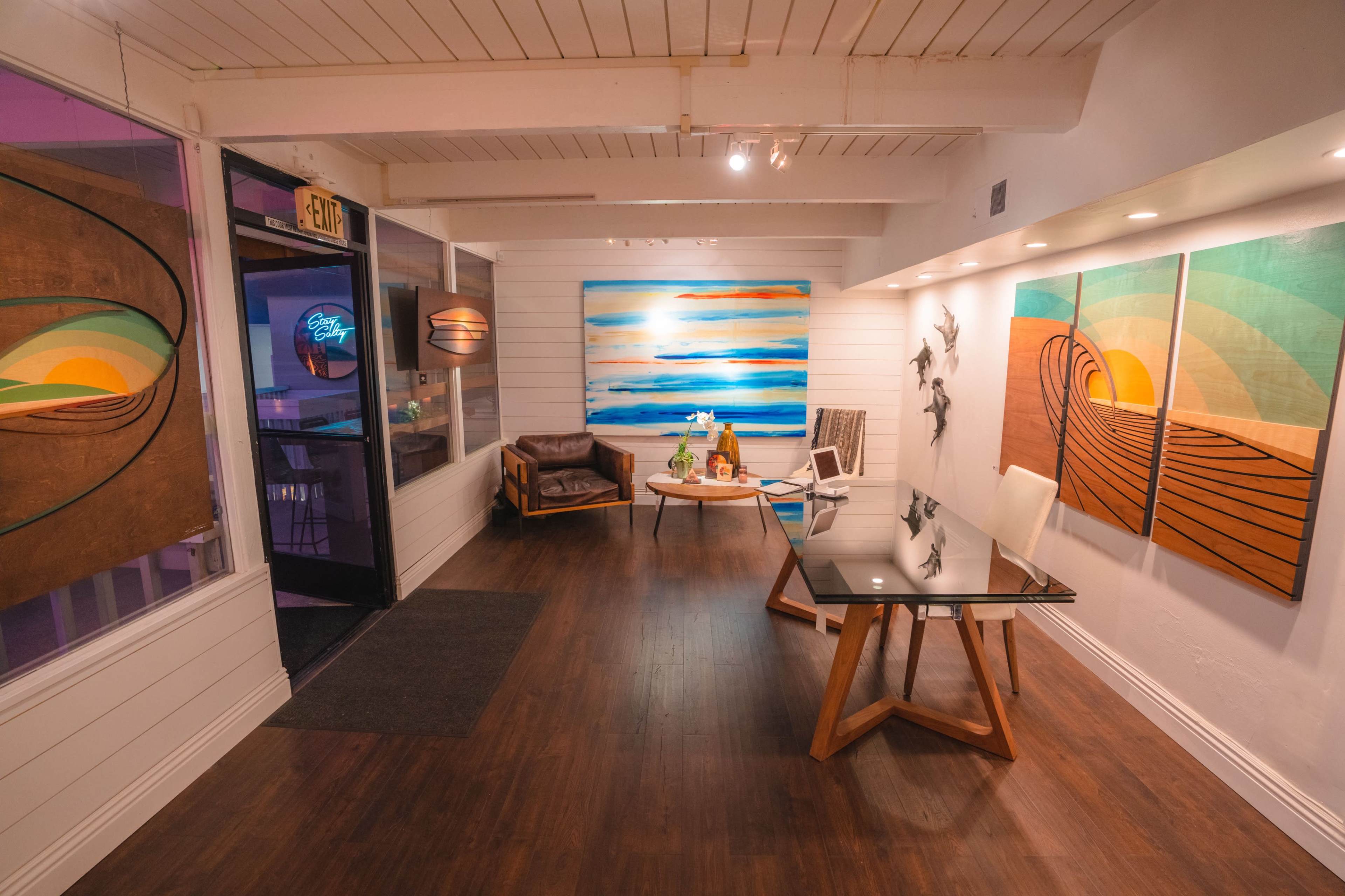 The interior of a gallery features colorful abstract paintings on the walls, a modern wooden table, and a cozy seating area with a leather chair.
