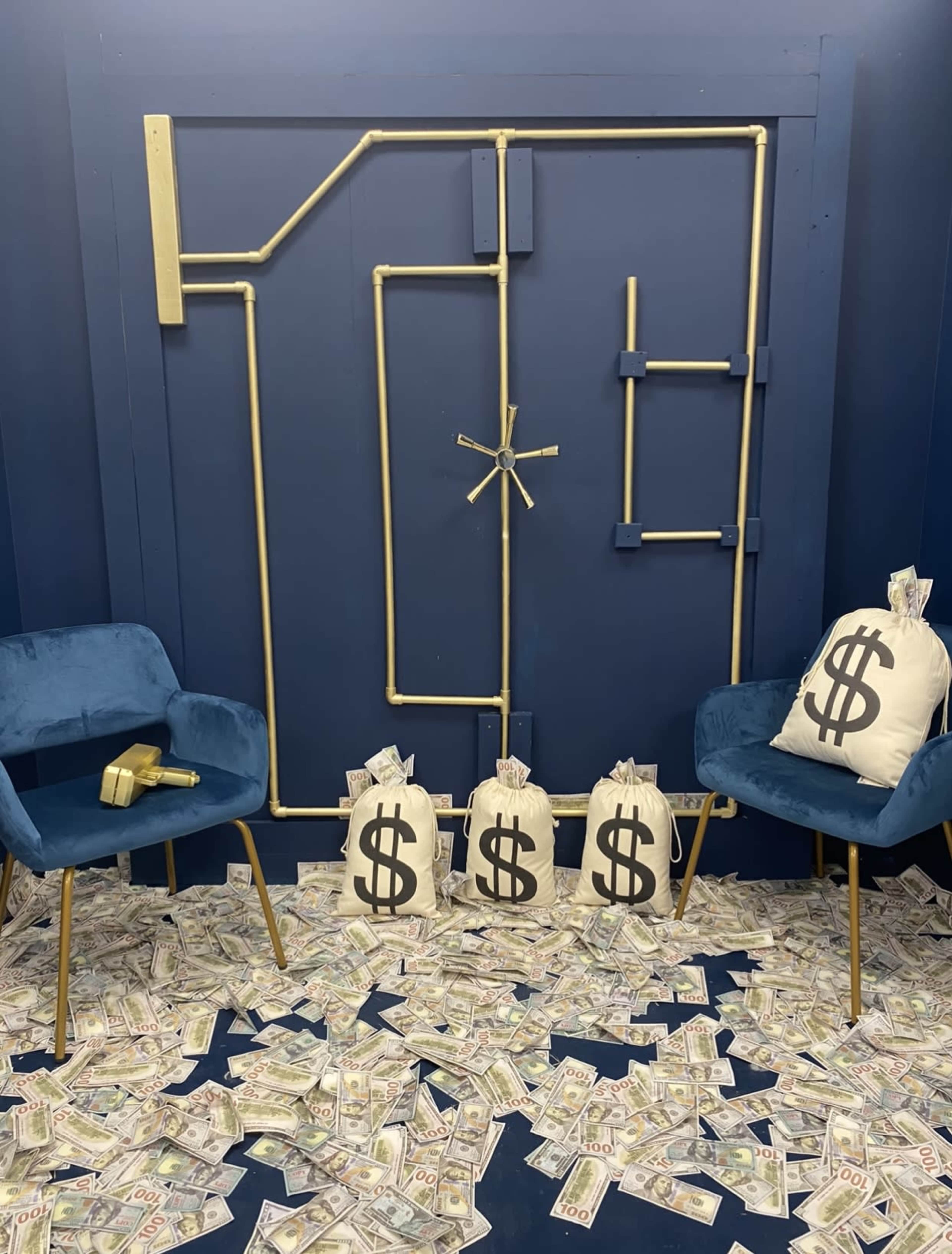 A blue room with a gold pipe design resembling a vault door, surrounded by stacks of fake money scattered on the floor and bags marked with dollar signs.
