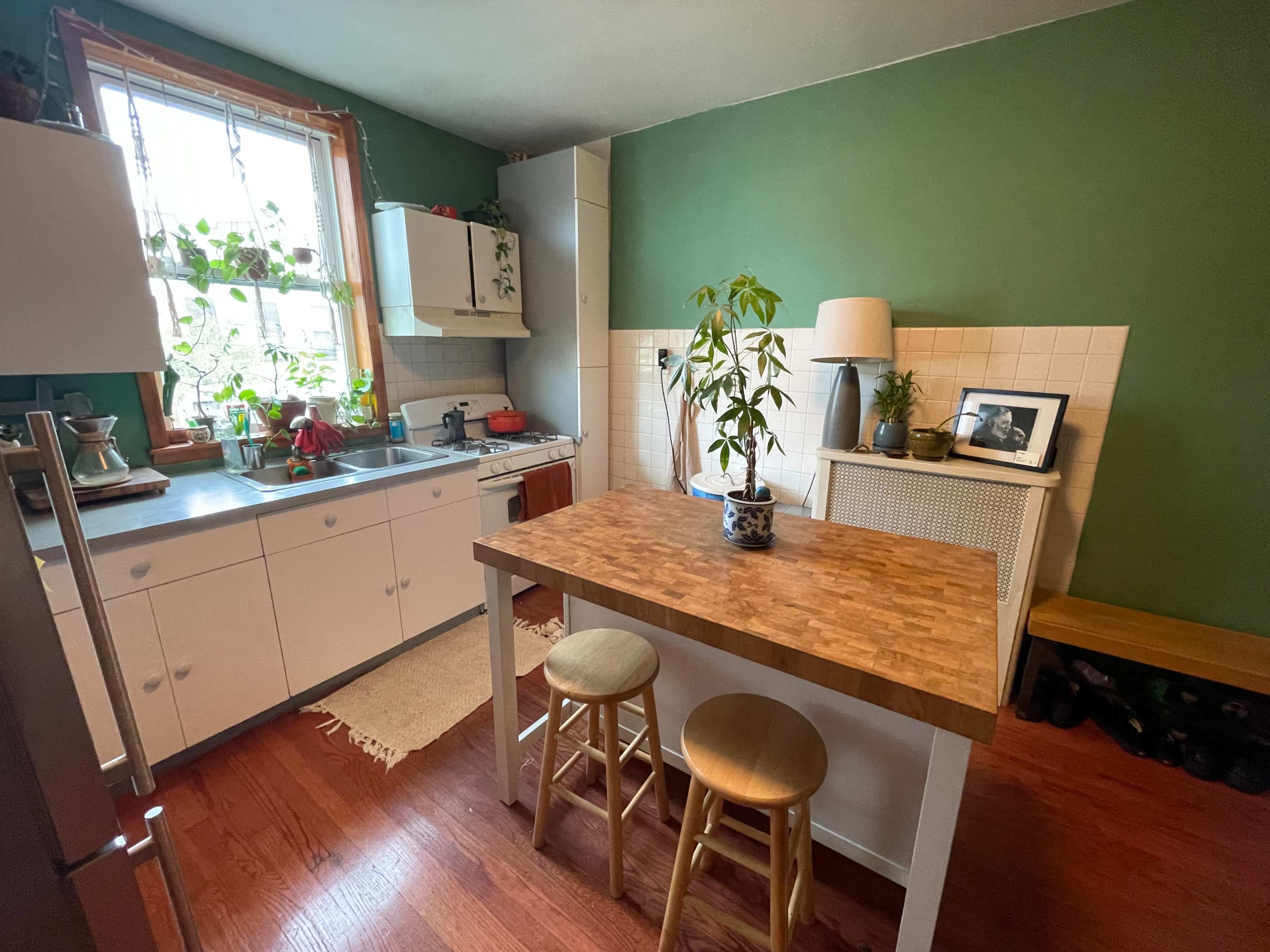 A kitchen with green walls, a wooden table, and stools, featuring plants and kitchen appliances arranged against the walls.
