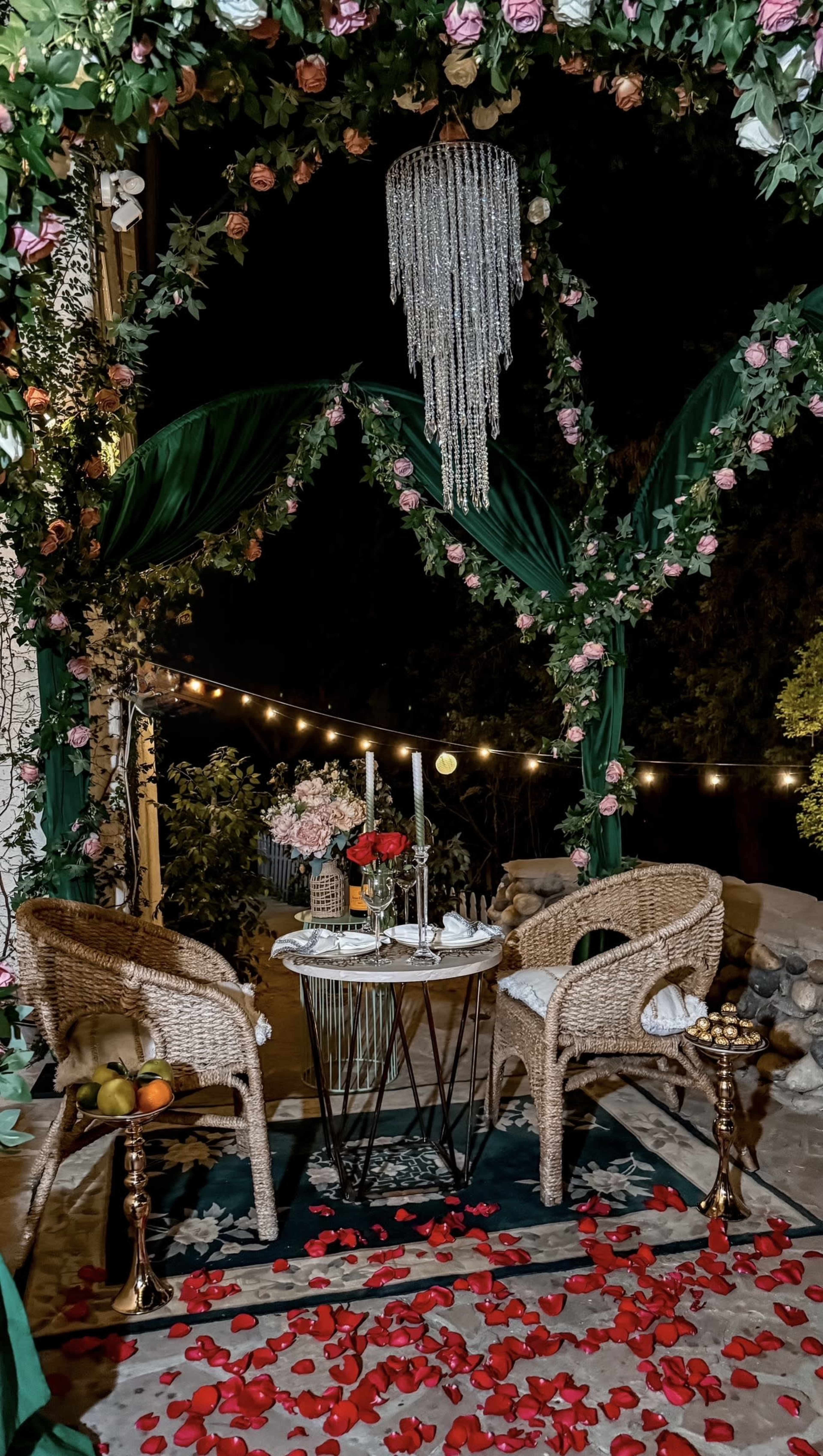 A cozy outdoor dining setup features two wicker chairs, a small round table adorned with a candle and flowers, surrounded by rose petals and draped with greenery and string lights.