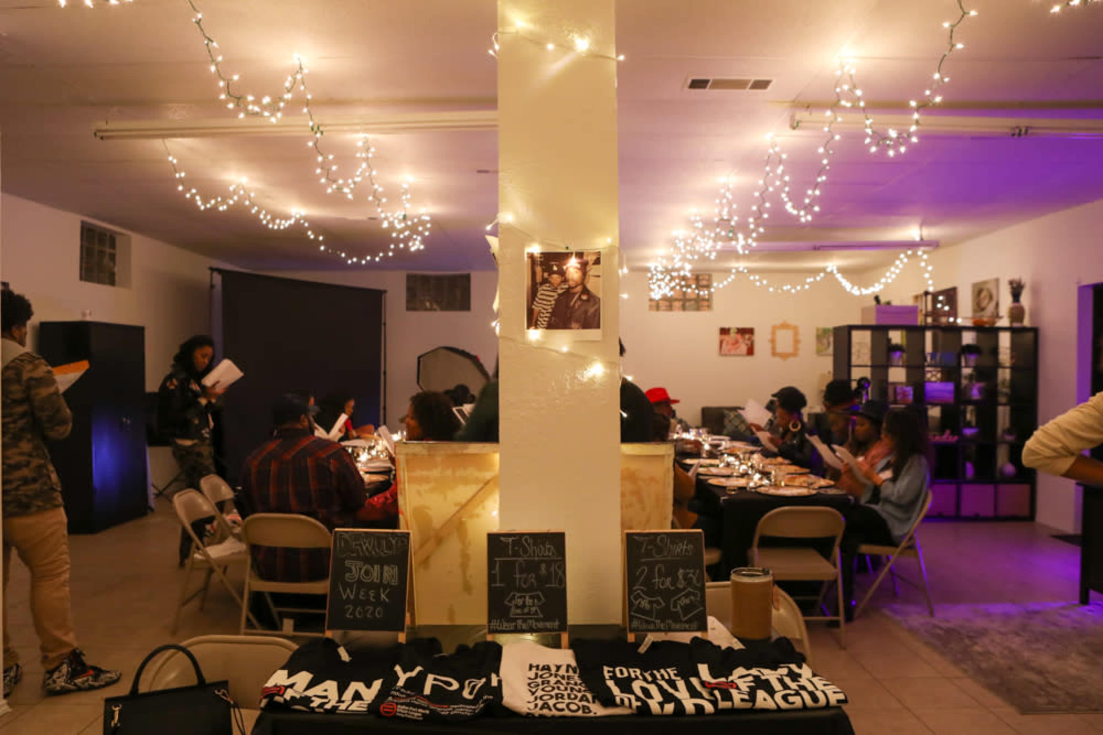 The image shows a dimly lit room with string lights, where a group of people is seated at tables engaged in conversation, while merchandise is displayed on a table in the foreground.