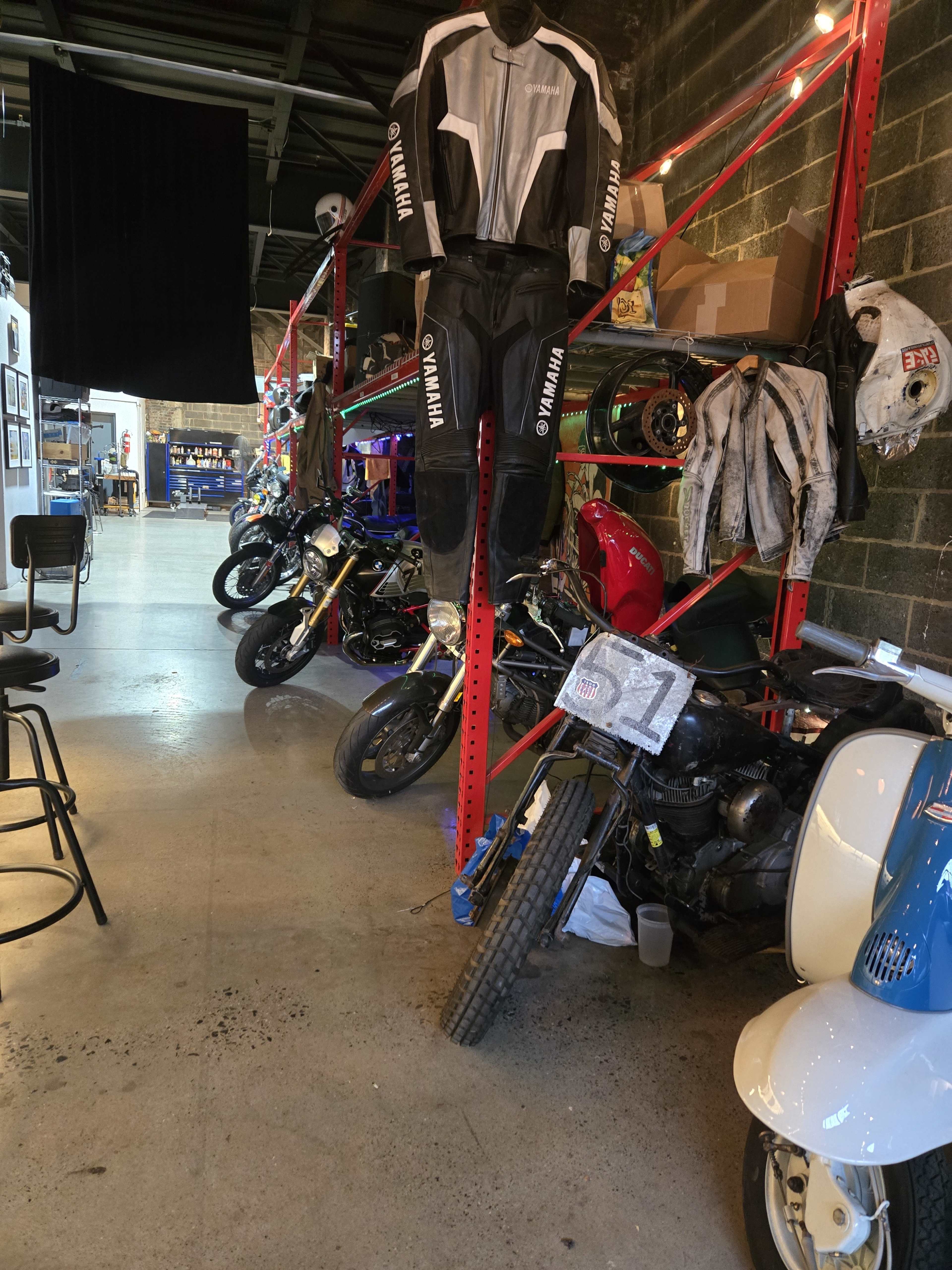 The image shows a garage filled with various motorcycles and race gear, including a Yamaha racing suit hanging on a rack.