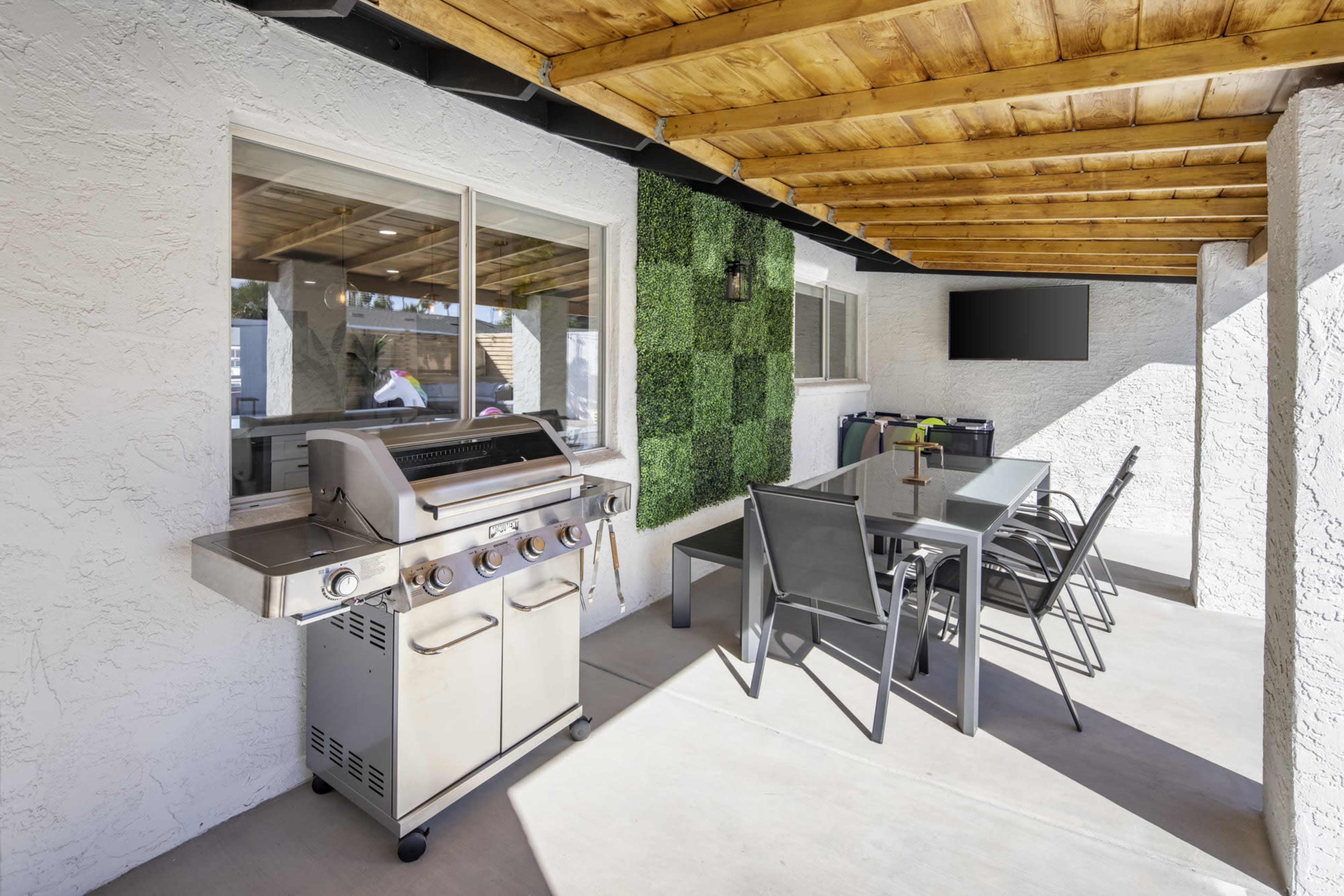 An outdoor patio area with a barbecue grill, a glass dining table, and metal chairs under a wooden ceiling.