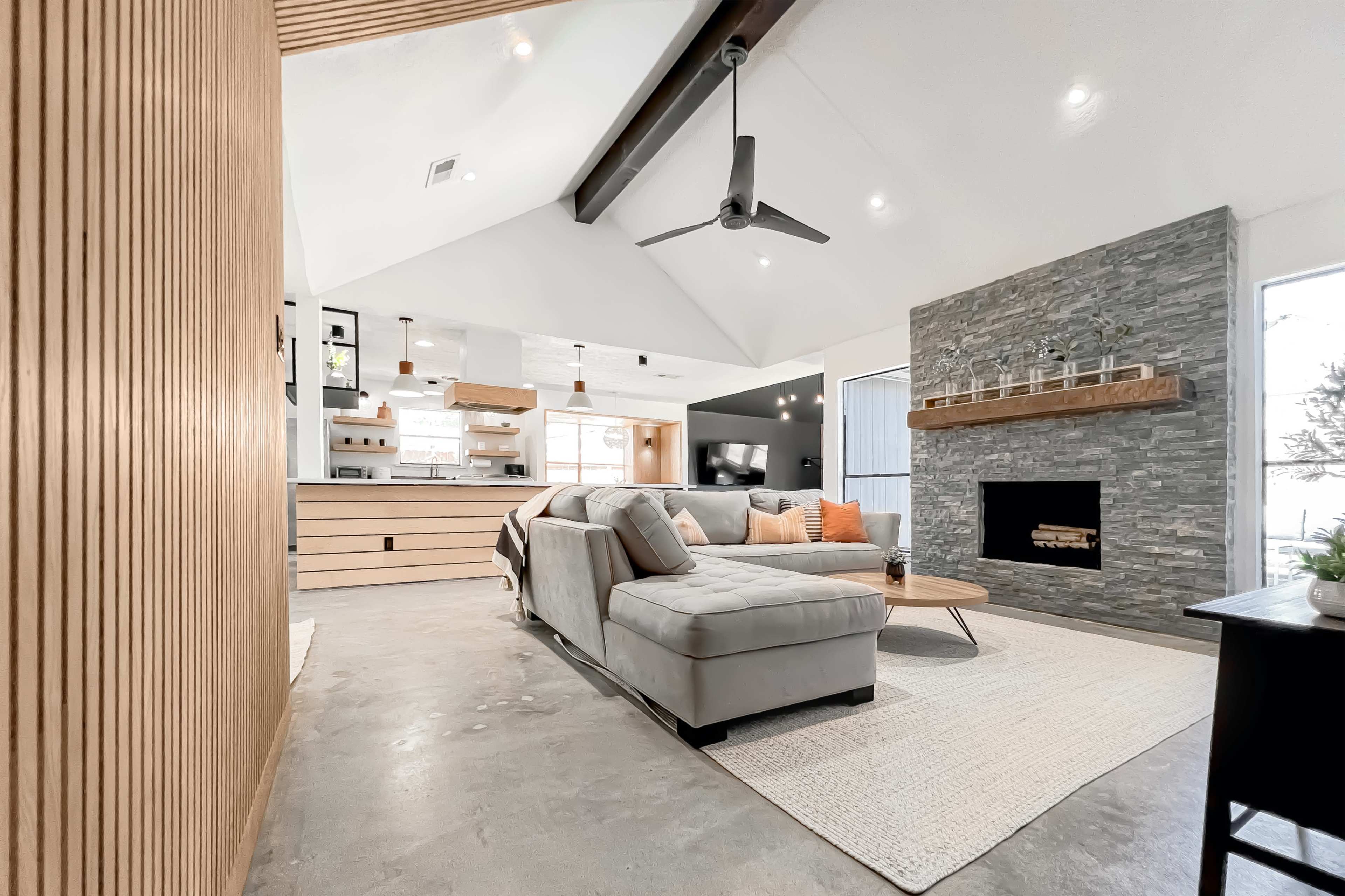 The photo shows a contemporary living room featuring a sectional sofa, a stone fireplace, and a kitchen area in an open layout with wooden cabinetry.