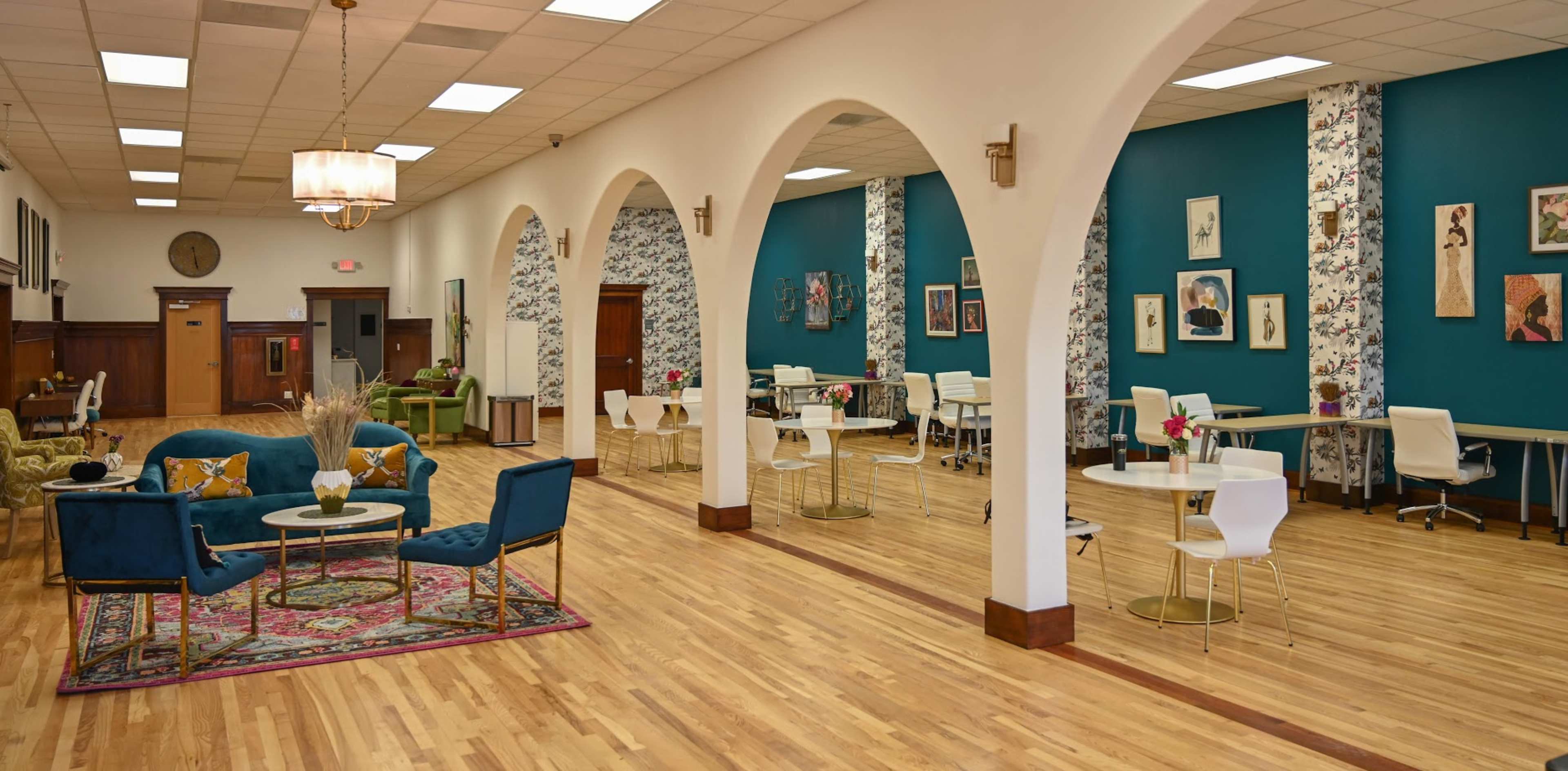 A spacious, well-lit interior featuring wooden flooring, arched doorways, and a mix of seating areas with tables and chairs against a backdrop of decorative walls.