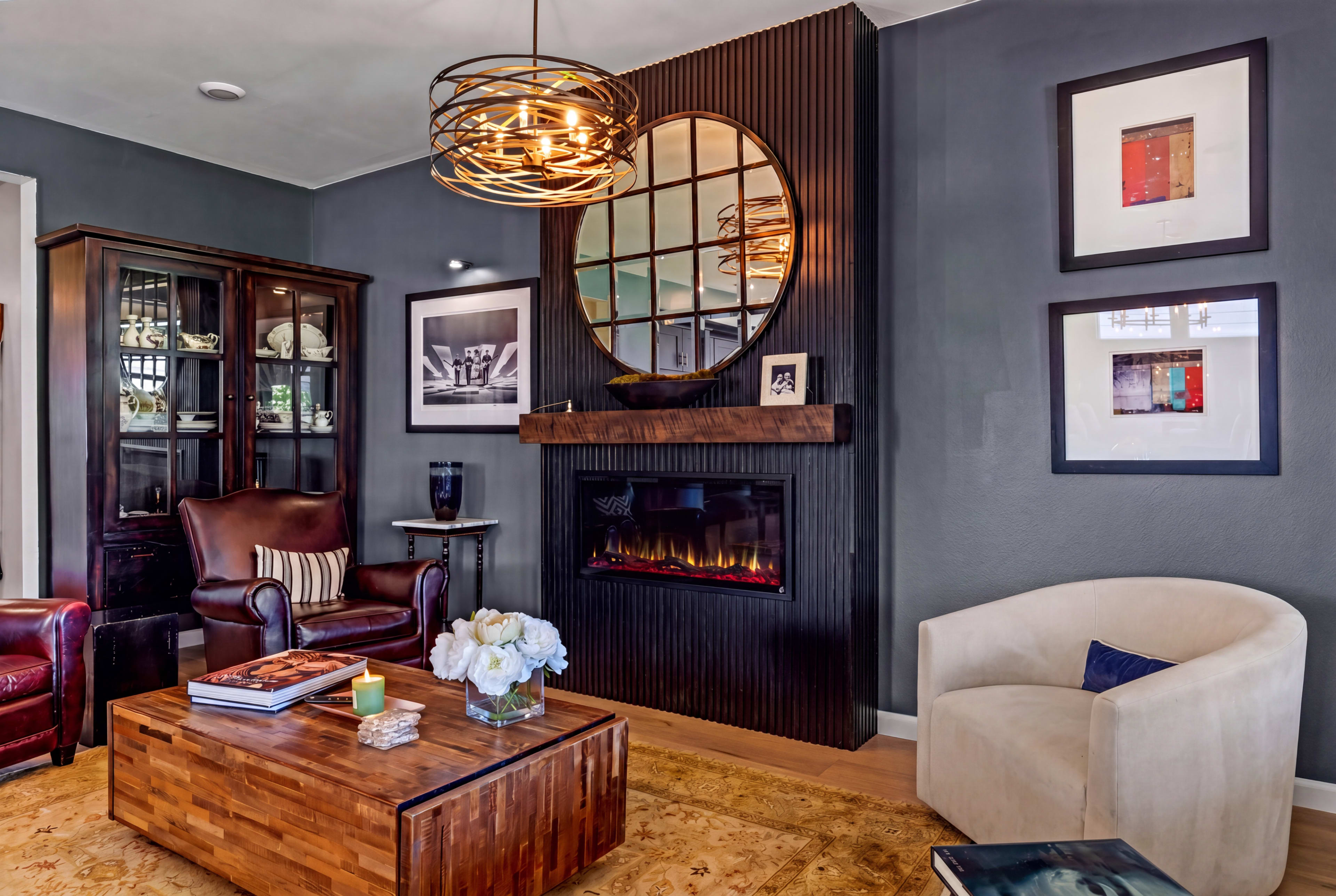 The image depicts a cozy living room featuring a modern fireplace, a round mirror, and a mix of stylish furniture arranged around a wooden coffee table.