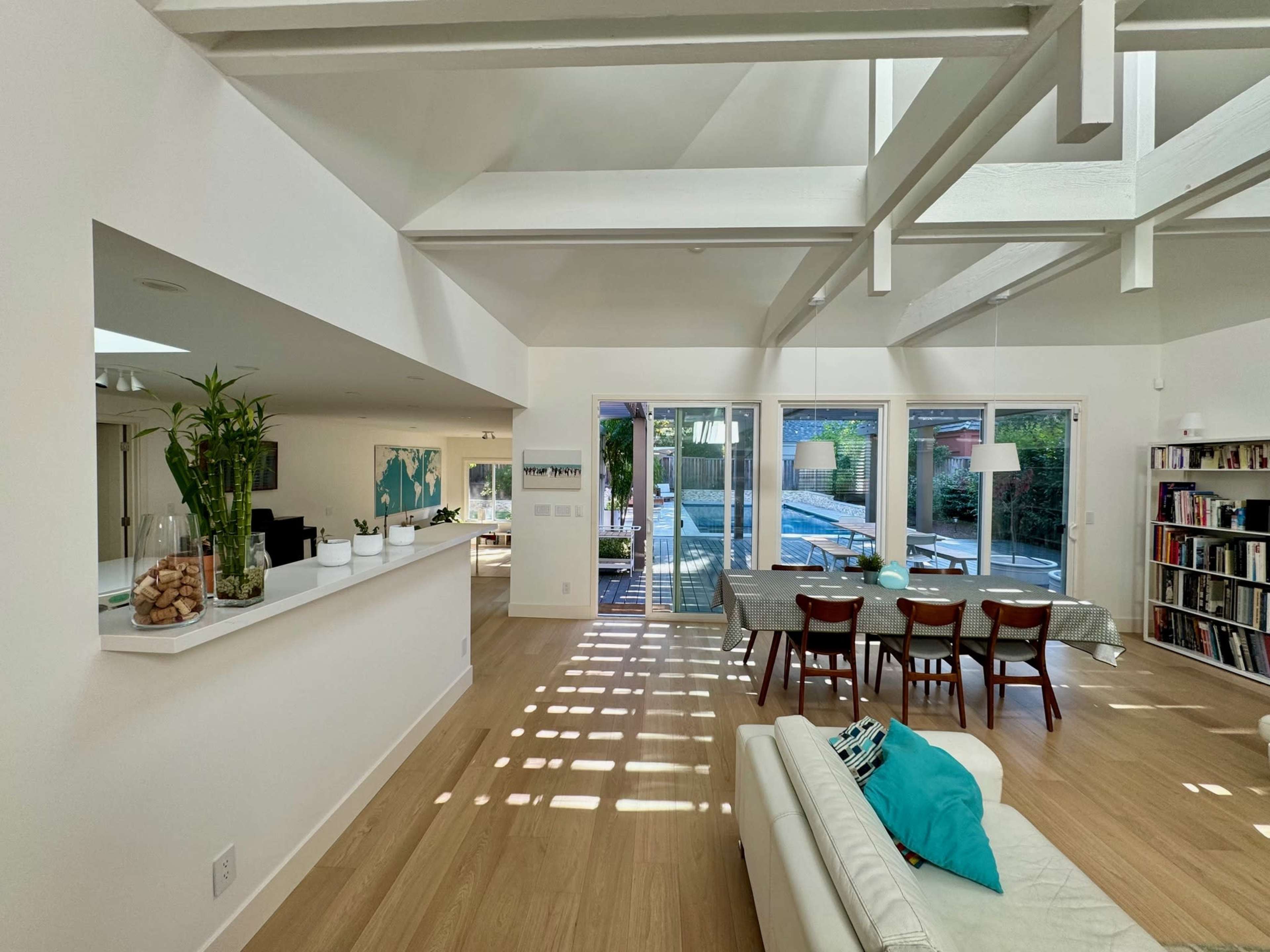 The image shows a bright interior space featuring a kitchen, dining area, and living room with a view of a pool and outdoor area through large glass doors.