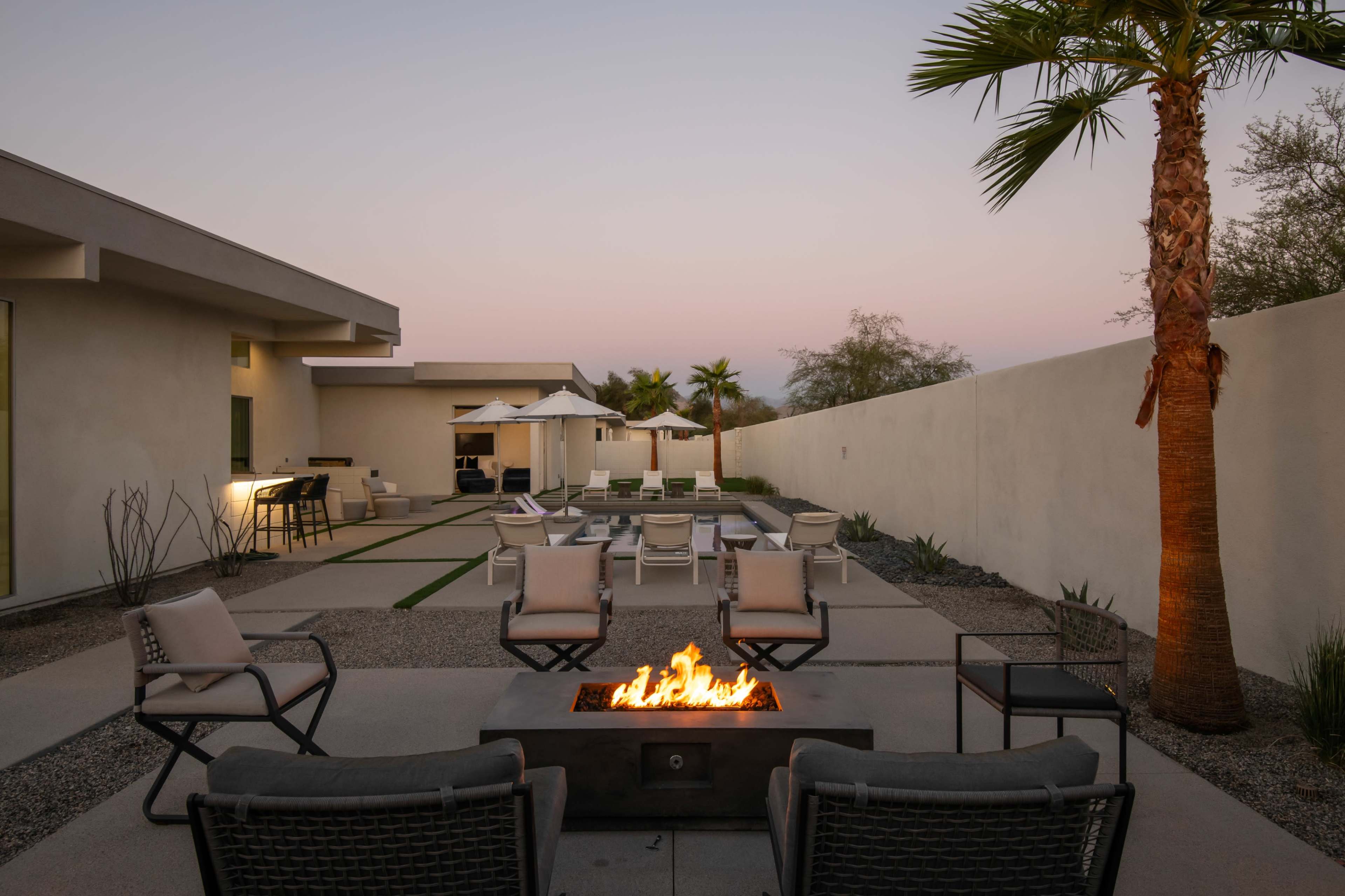 A modern outdoor patio features a fire pit surrounded by seating, with lounge chairs and umbrellas arranged in a spacious layout.