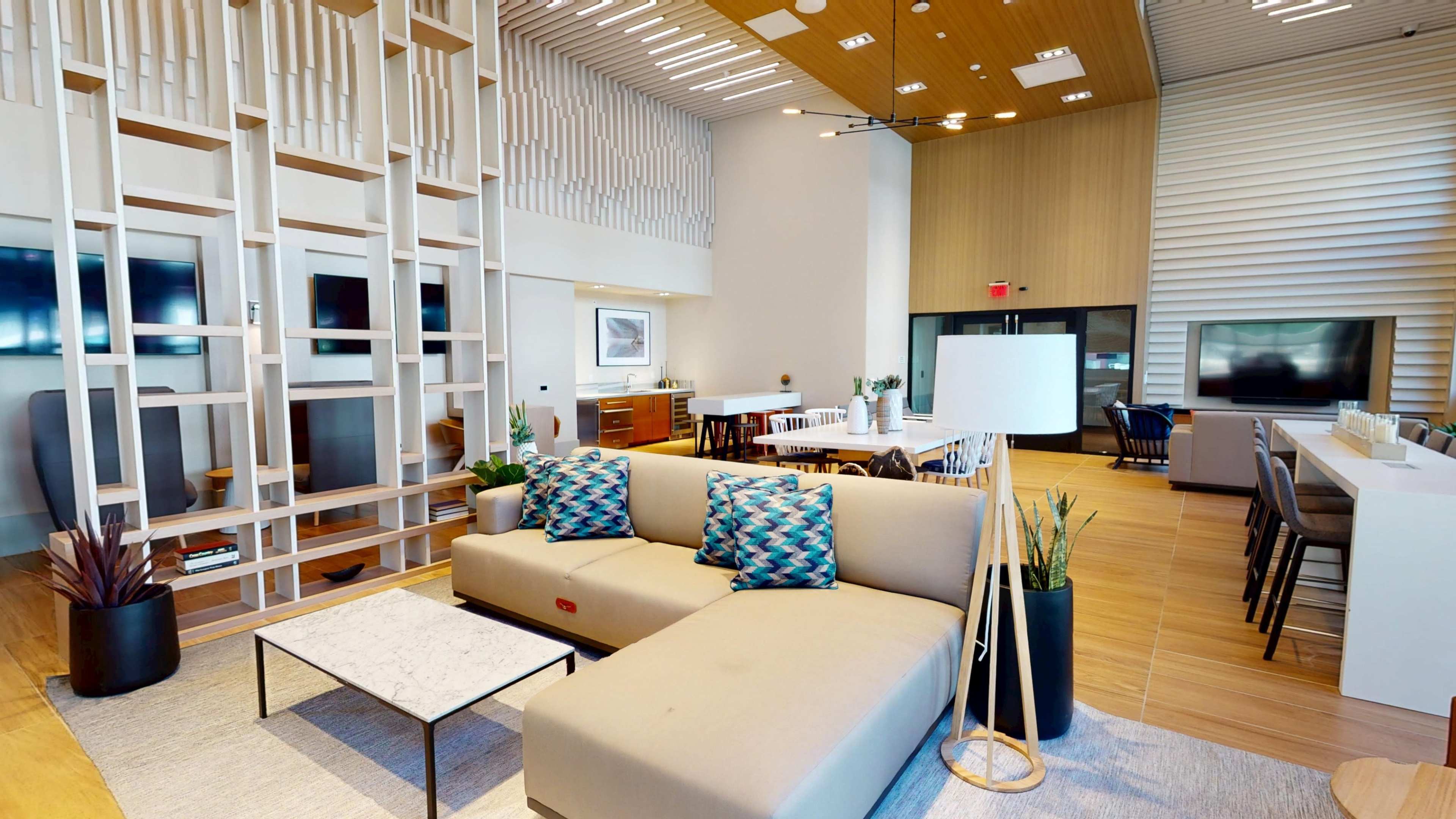 The image shows a modern, open-concept living space featuring a beige sofa with patterned cushions, a coffee table, and a dining area with multiple chairs and tables against a backdrop of wooden panels and large windows.