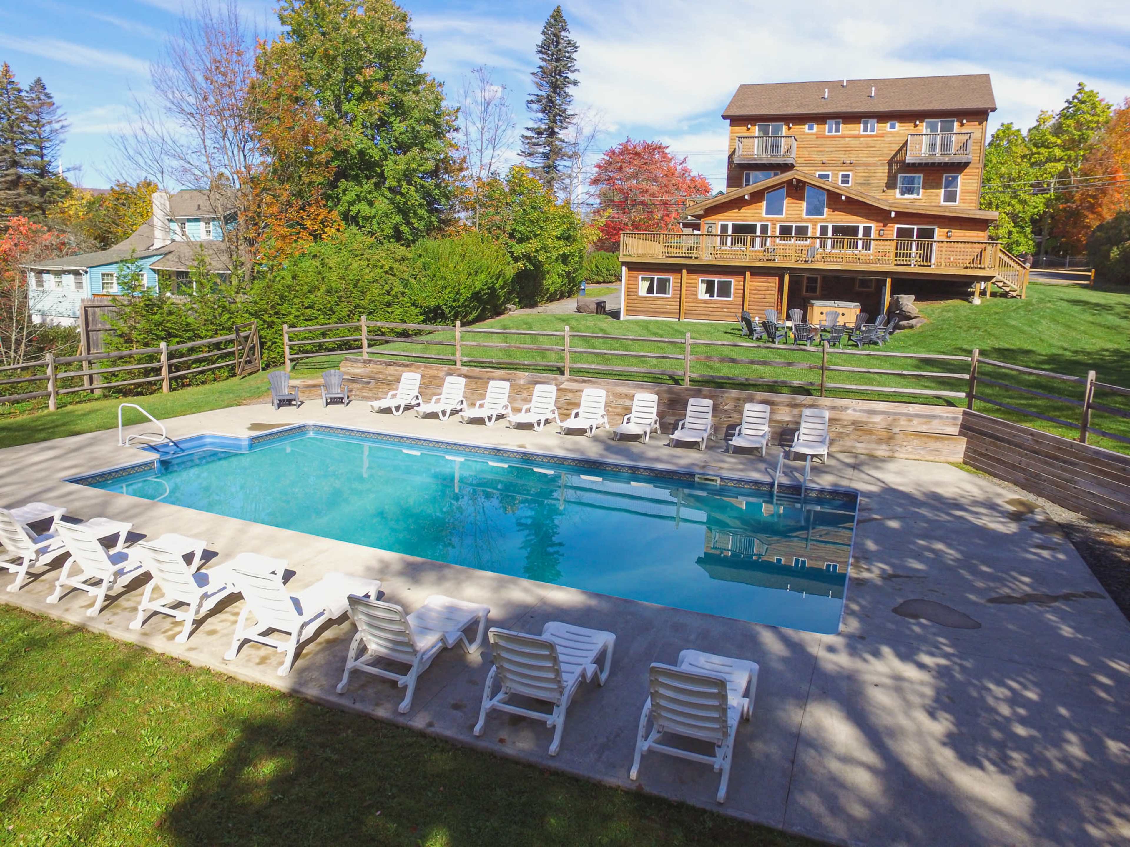 A large outdoor swimming pool is surrounded by lounge chairs, with a multi-story wooden building and trees in the background.