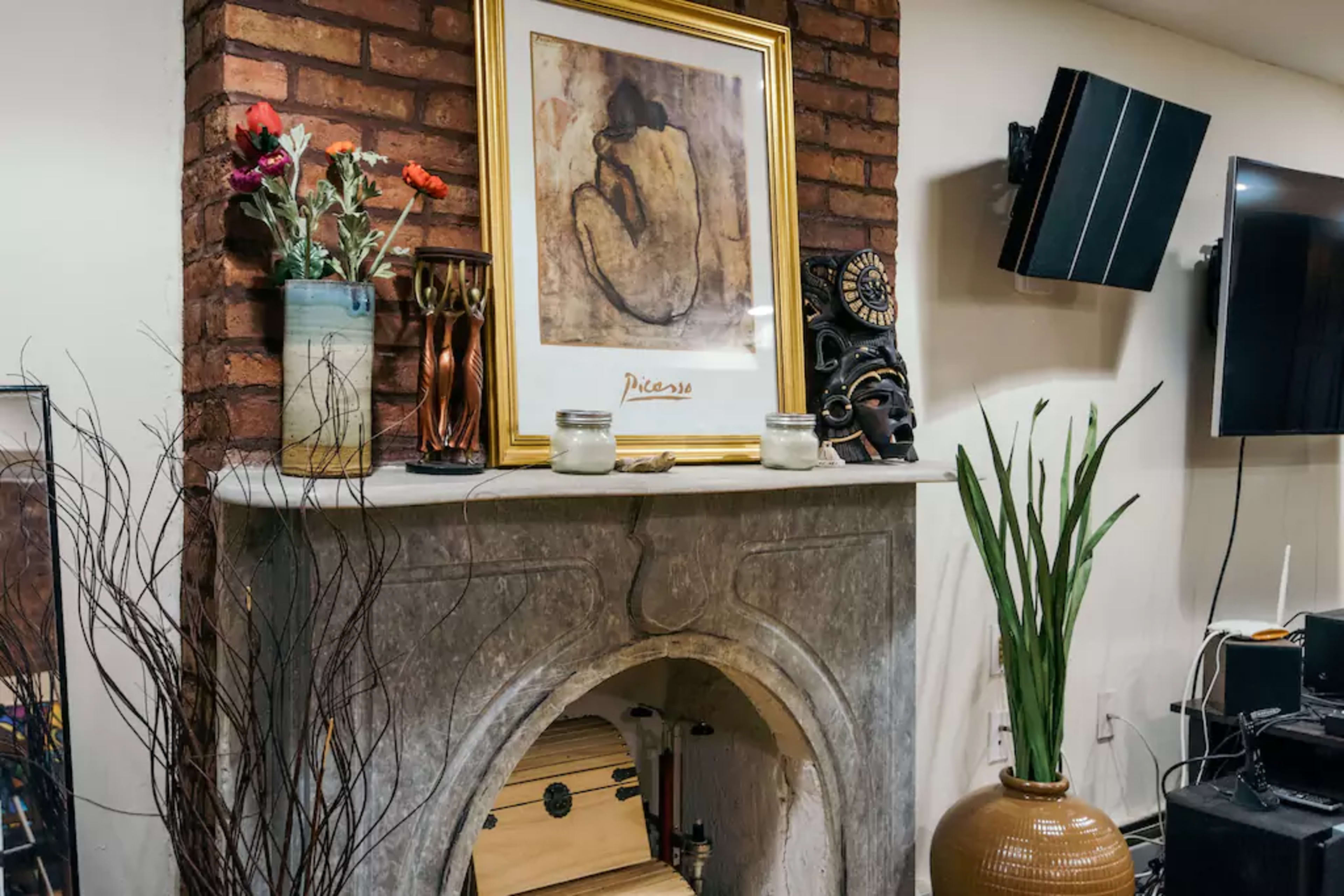 A decorative fireplace with a framed artwork above it, surrounded by various ornaments, including flowers and a plant, while a television and speakers are visible on the opposite wall.