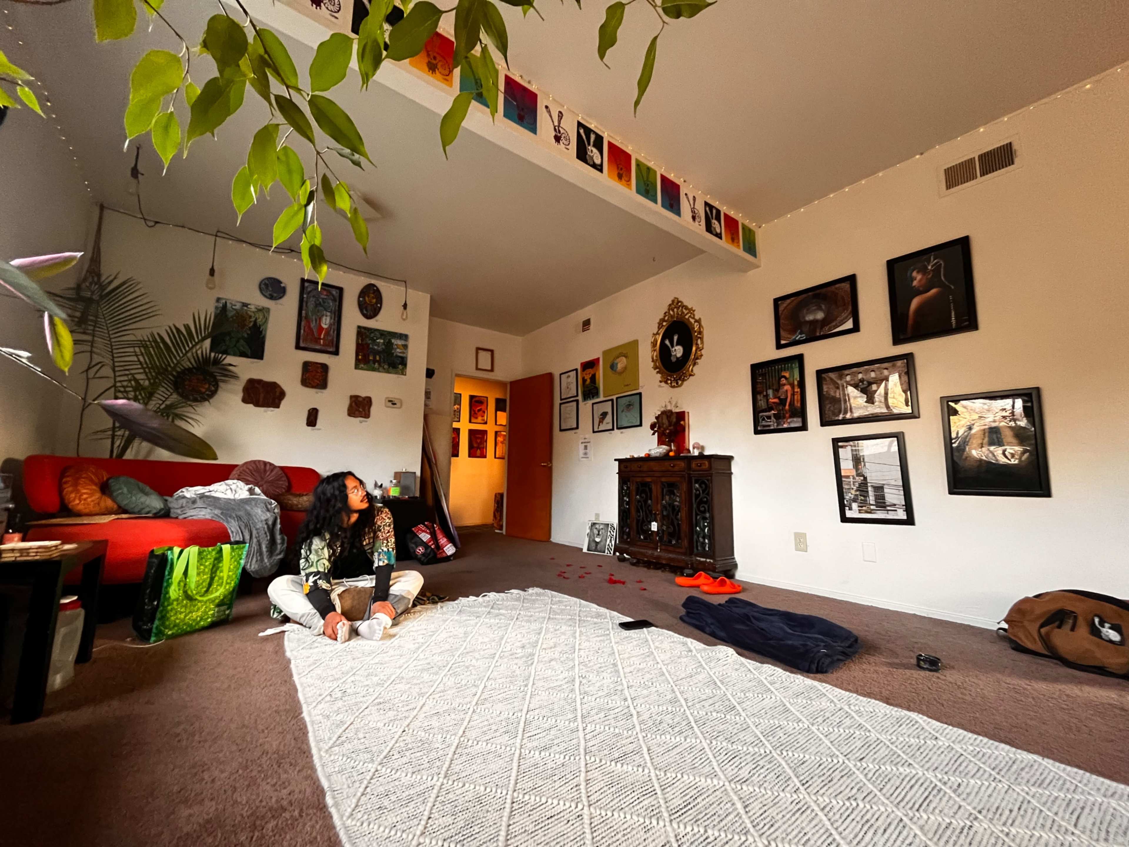 A person sits on the floor of a living room with a textured rug, surrounded by various artworks and decorations on the walls.