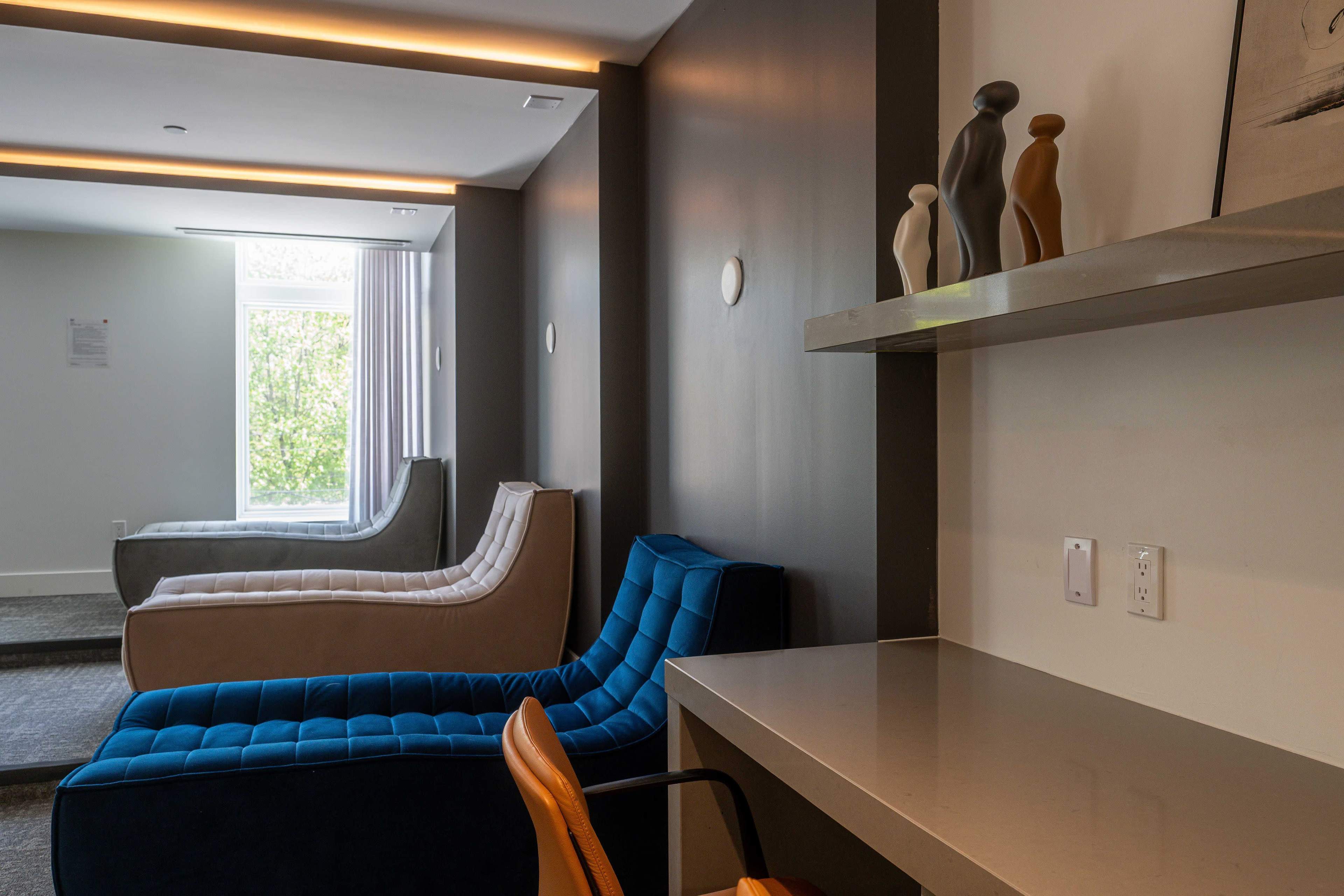 The image shows a modern lounge area with two curved seating structures, a blue armchair, and a shelf displaying decorative objects.