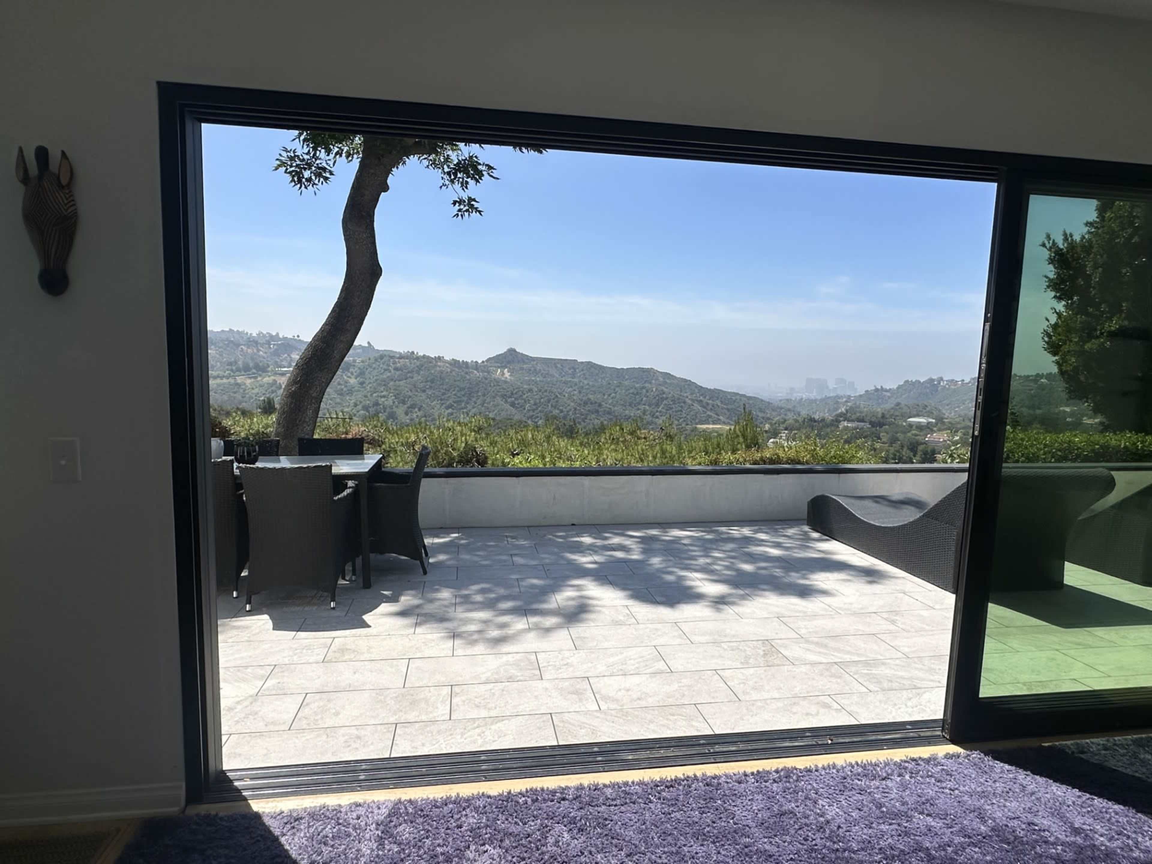 The image shows an open glass door leading to a patio with a table and chairs, overlooking a hilly landscape.