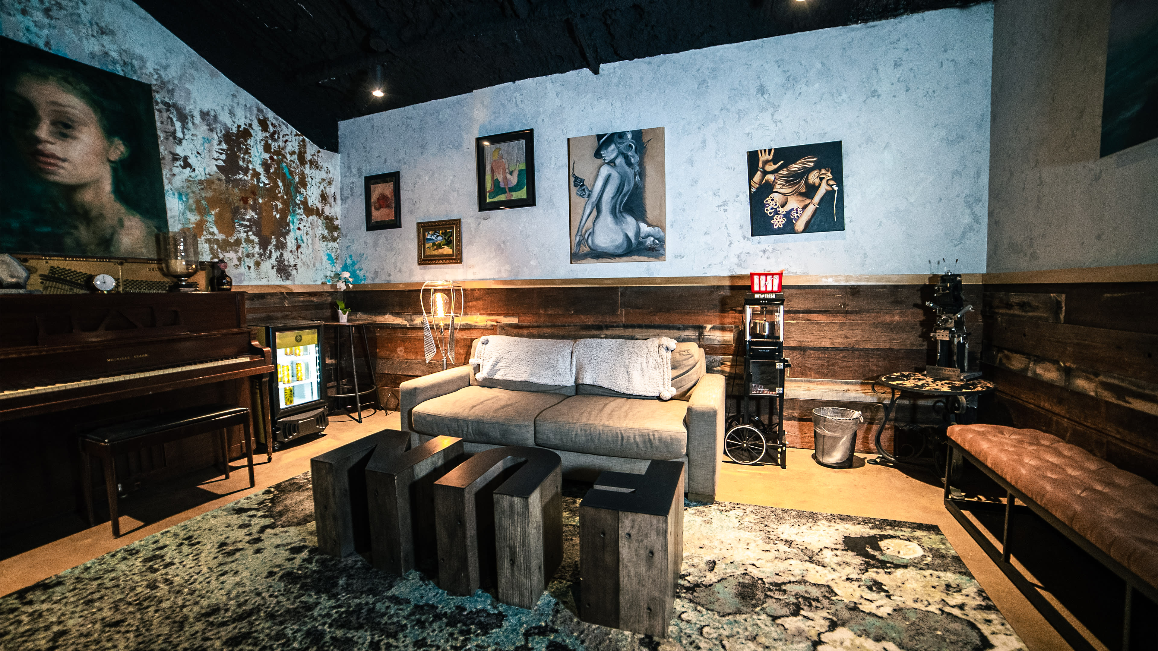 A cozy room with a brown sofa and wooden coffee table, surrounded by various artworks on the walls and a piano in the corner.