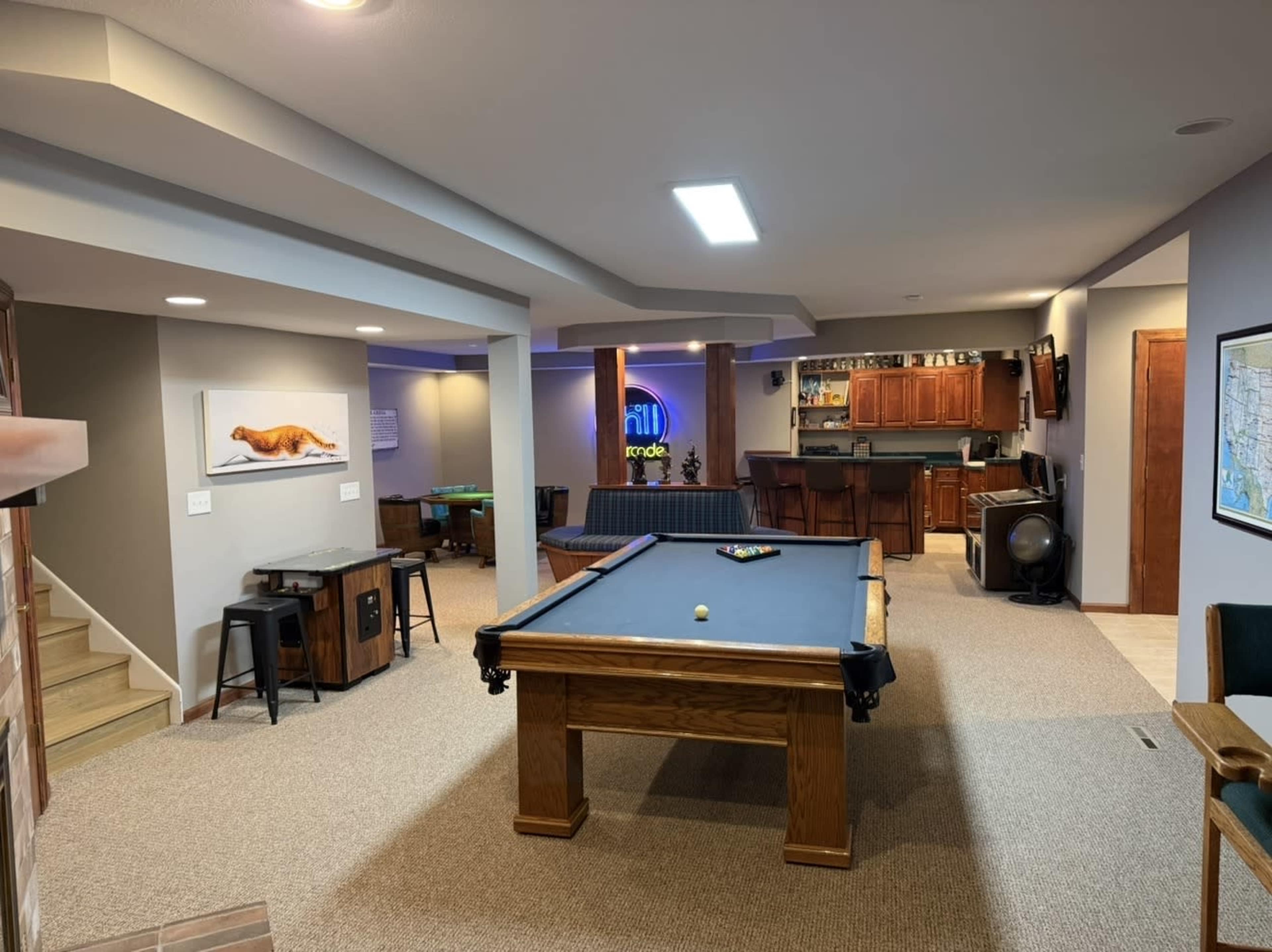 The image shows a spacious basement game room featuring a pool table, seating areas, and a bar counter in the background.