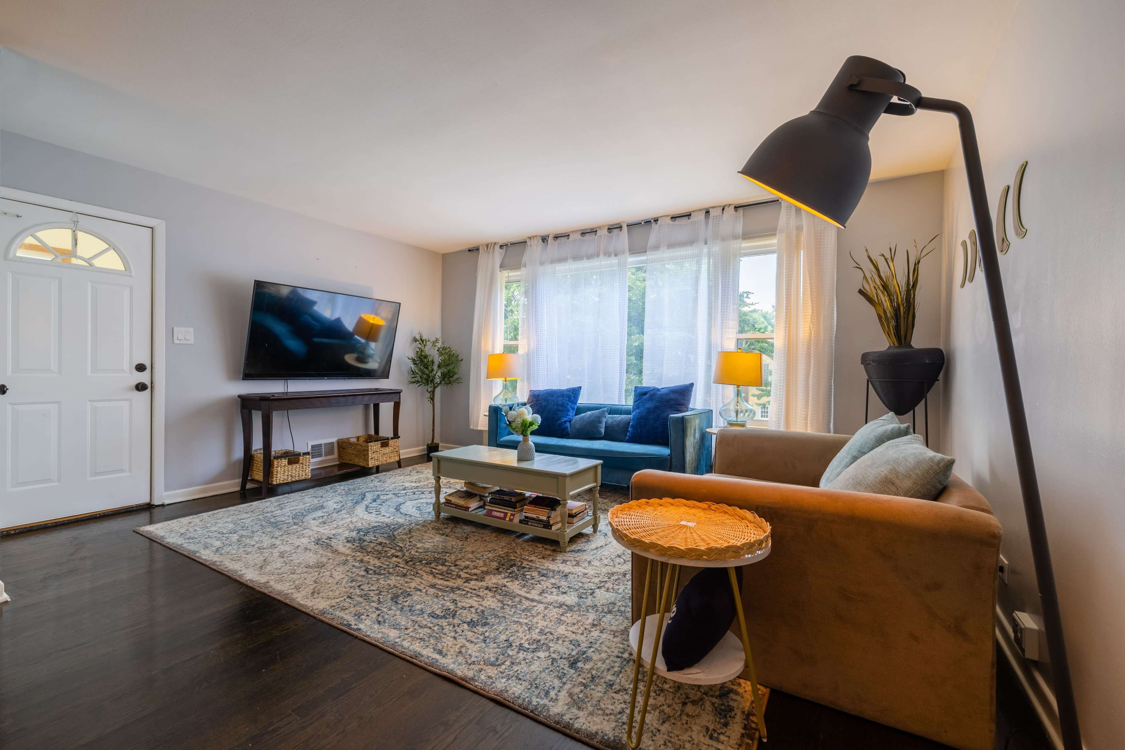 A modern living room features a sofa, coffee table, and a large television against a light-colored wall with natural light streaming through sheer curtains.