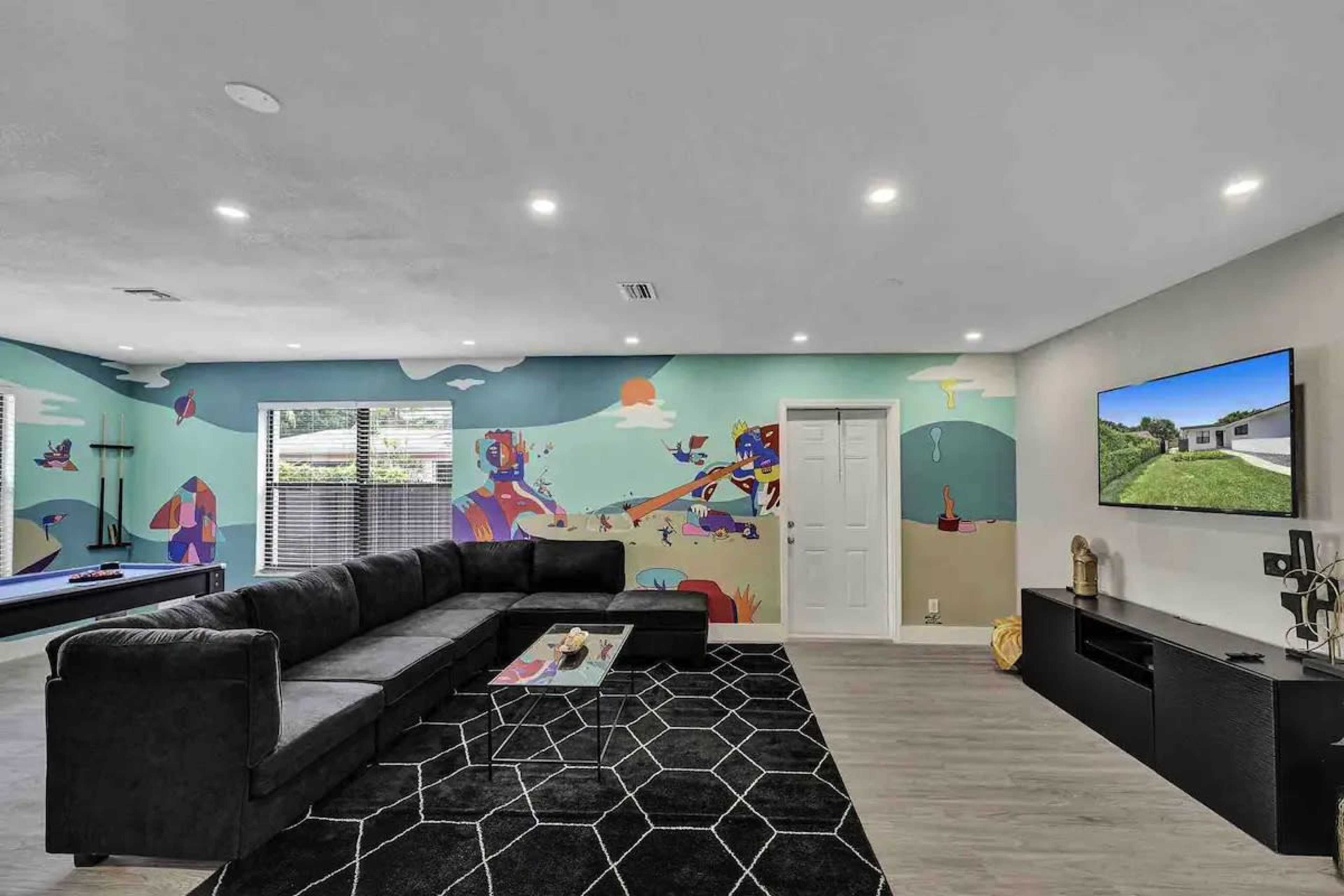 The living room features a large sectional sofa, a colorful mural on the wall, a TV mounted on a side wall, and a modern black rug on the floor.