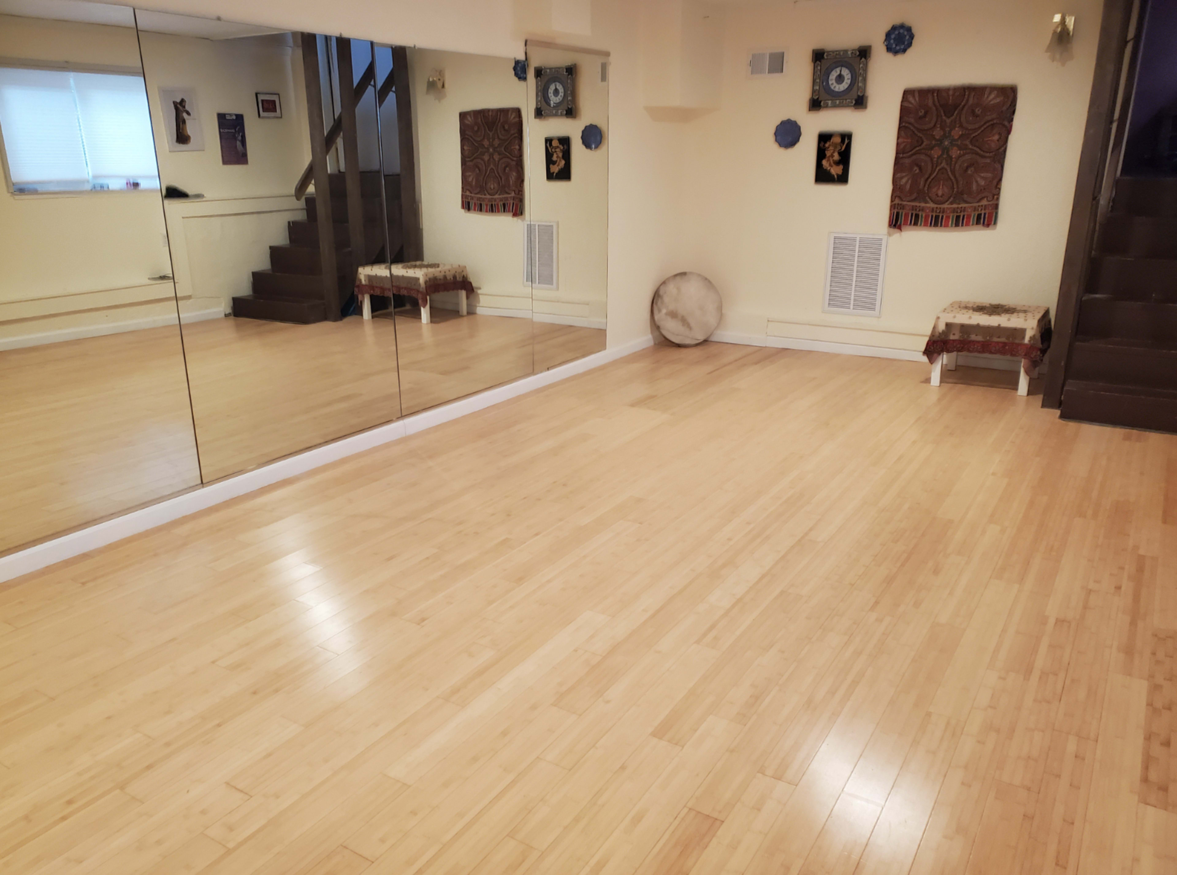 The image shows a spacious room with bamboo flooring, a large mirror on one wall, and decorative wall hangings.