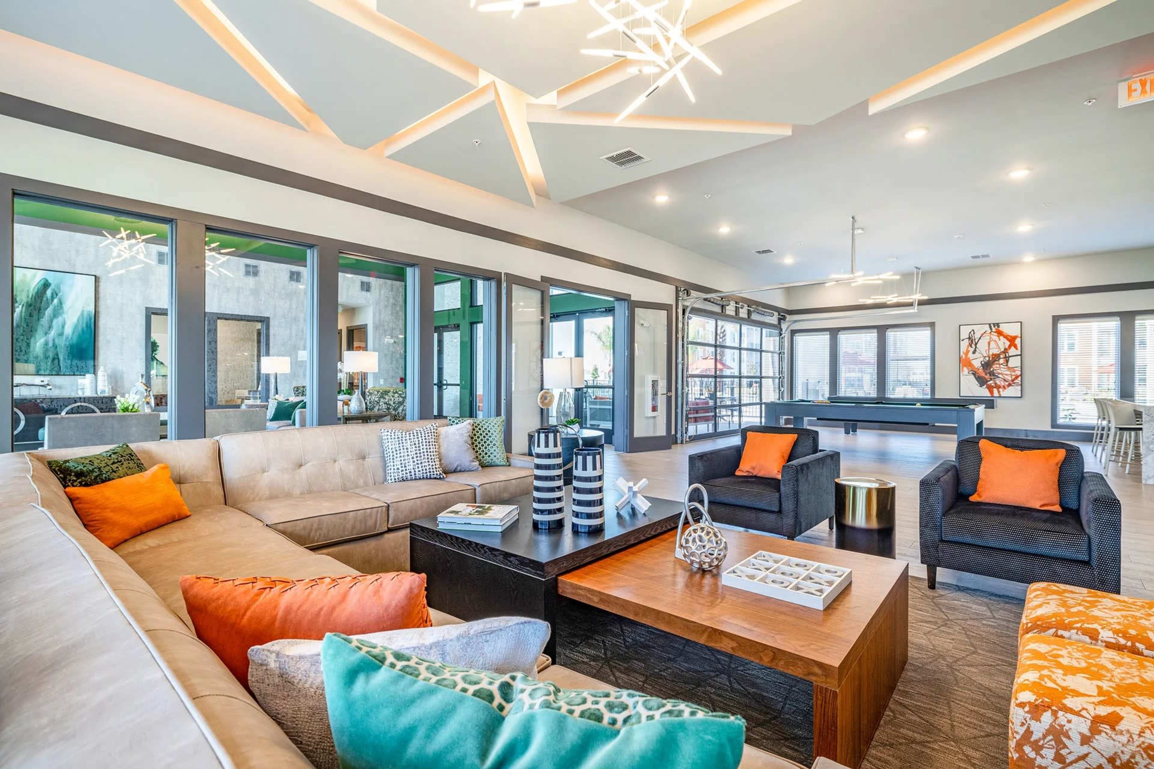 The image shows a stylish modern lounge area with a large sectional couch, two armchairs, a wooden coffee table, and large windows letting in natural light.