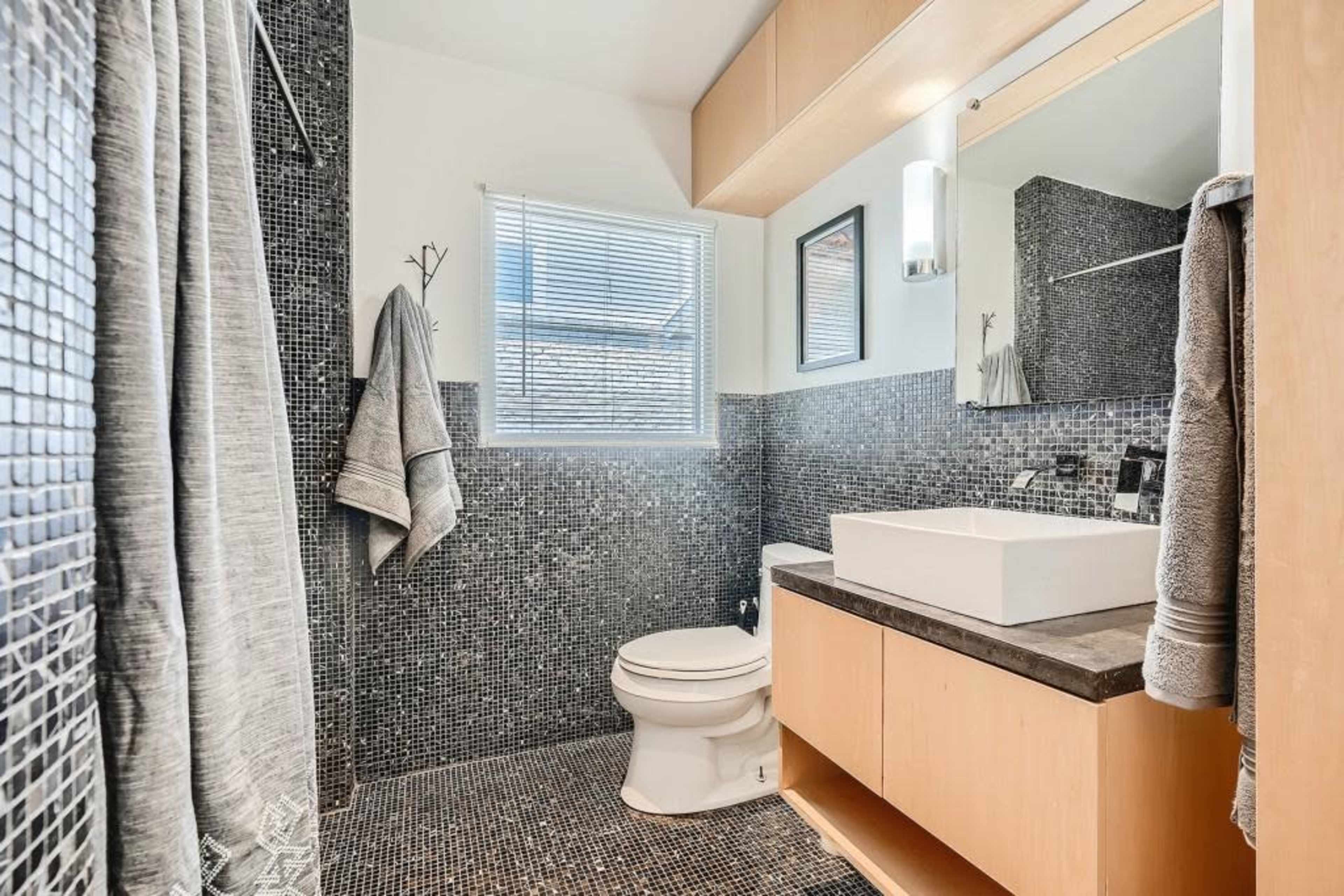 A modern bathroom featuring black mosaic tile walls, a white vessel sink on a wooden countertop, a toilet, and a shower with a gray curtain.