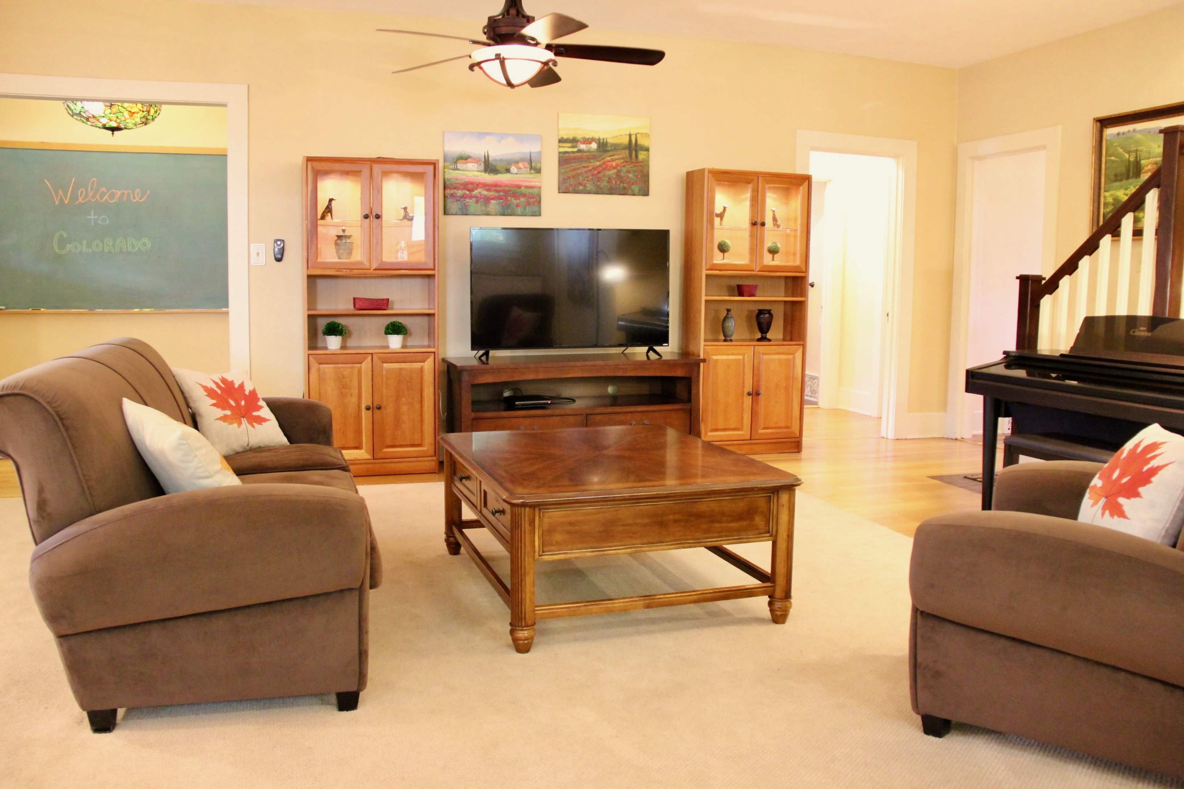 A living room with two brown sofas, a coffee table, a television on a stand, and a black piano, along with wooden shelves and a chalkboard reading "Welcome to Colorado."