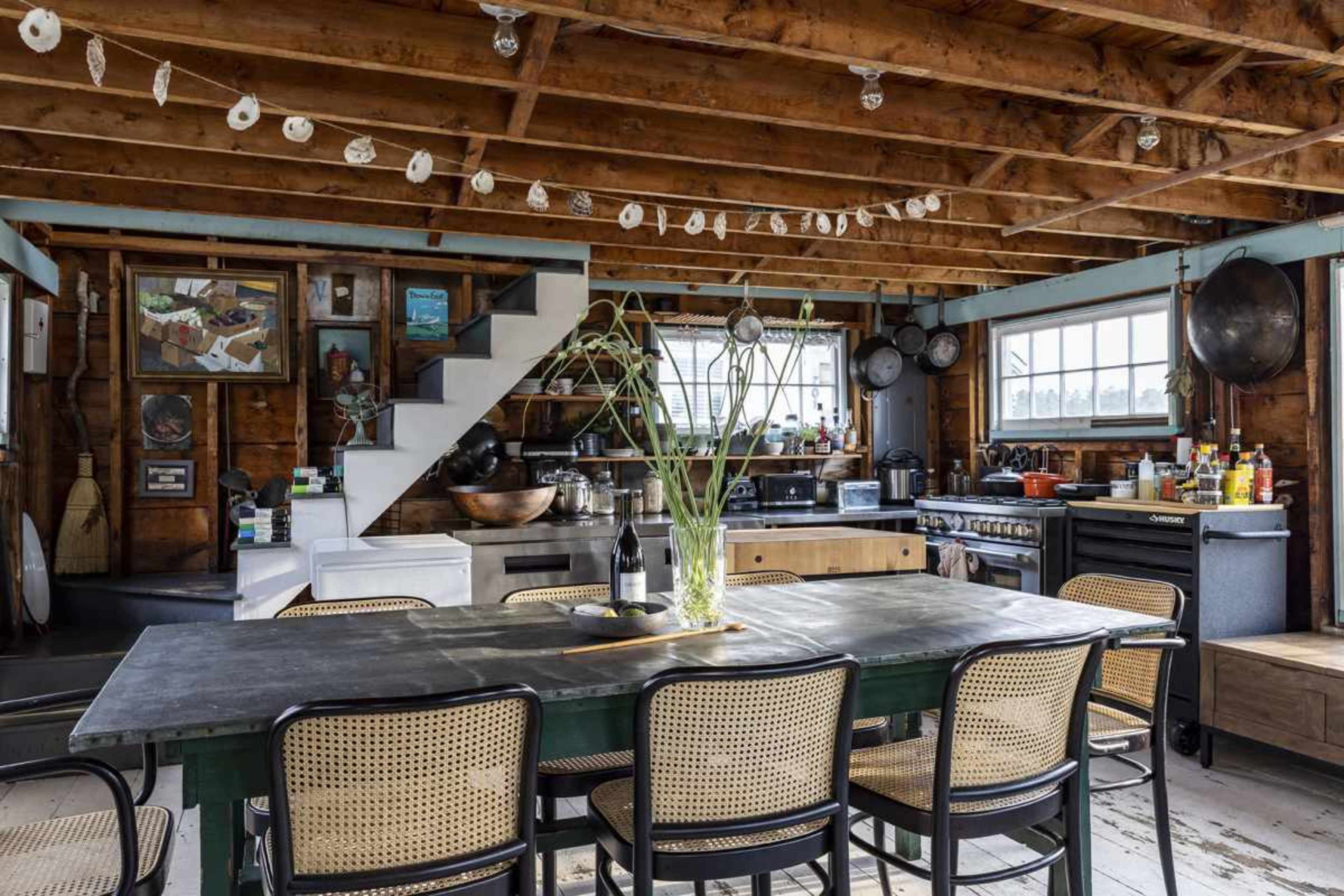 The image shows a rustic kitchen with a wooden ceiling, a dining table at the center, and a staircase leading to the upper level, surrounded by various kitchen appliances and decor.