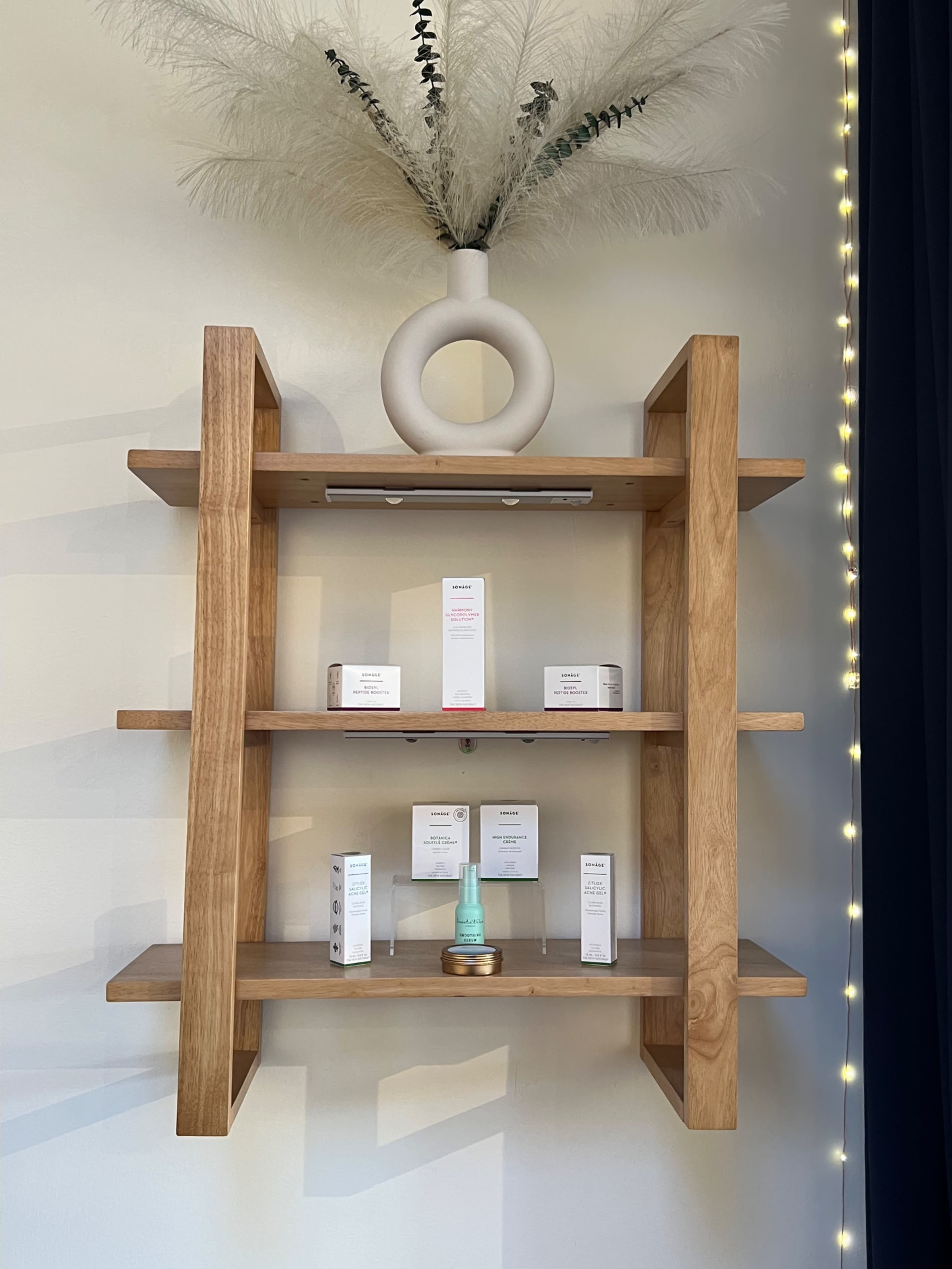 A wooden shelf displays various skincare products, with a decorative vase of pampas grass on top.