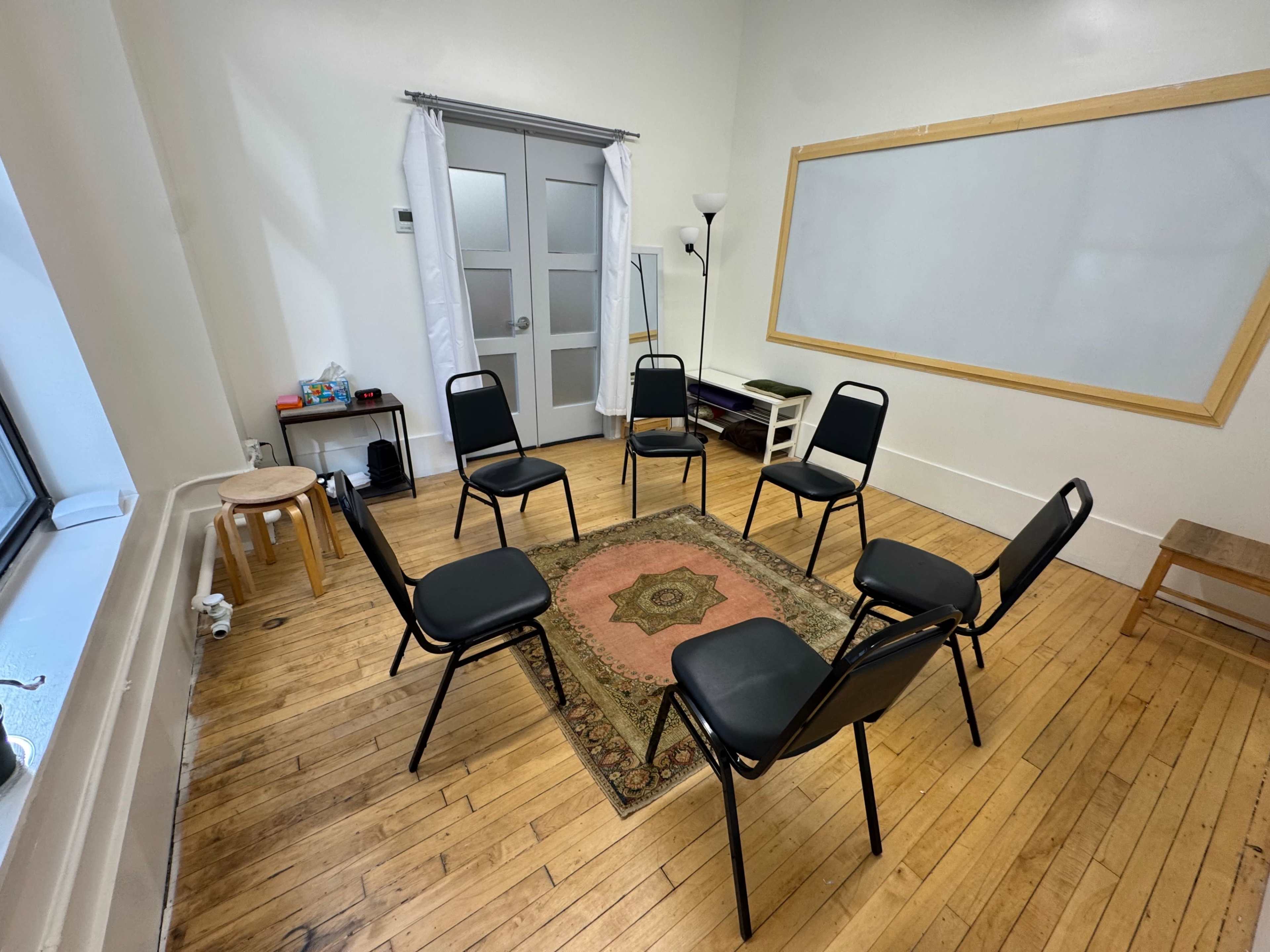 The image shows a small, well-lit room with six black chairs arranged in a circle on a wooden floor, surrounded by a few furnishings like a rug, a lamp, and a table against the wall.
