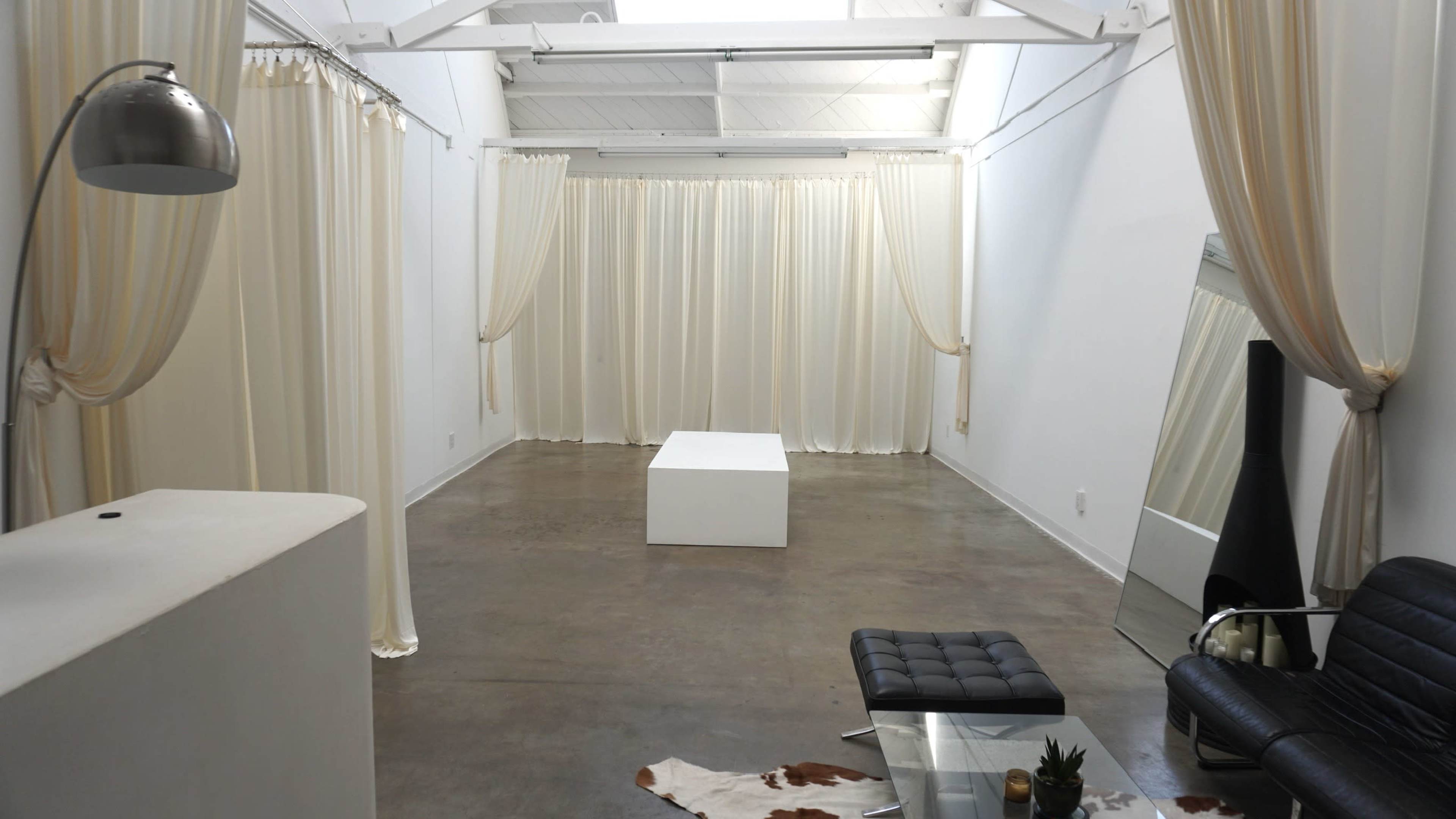 The image shows a spacious, minimally decorated room with white walls, a polished concrete floor, a black leather sofa, a white cube table, and sheer curtains hanging from the ceiling.