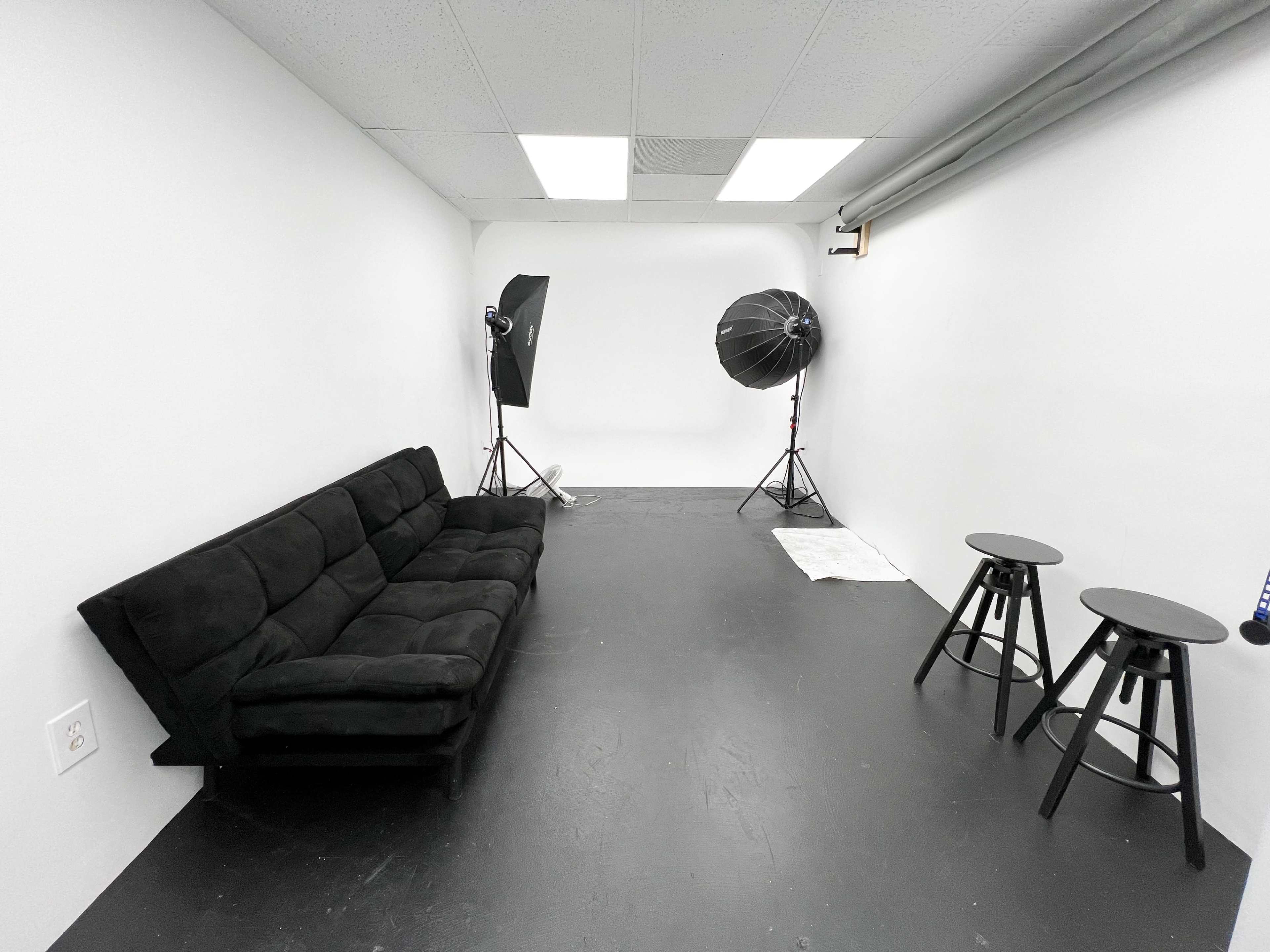 A minimalist studio space with a black sofa, two black stools, and lighting equipment set against white walls.