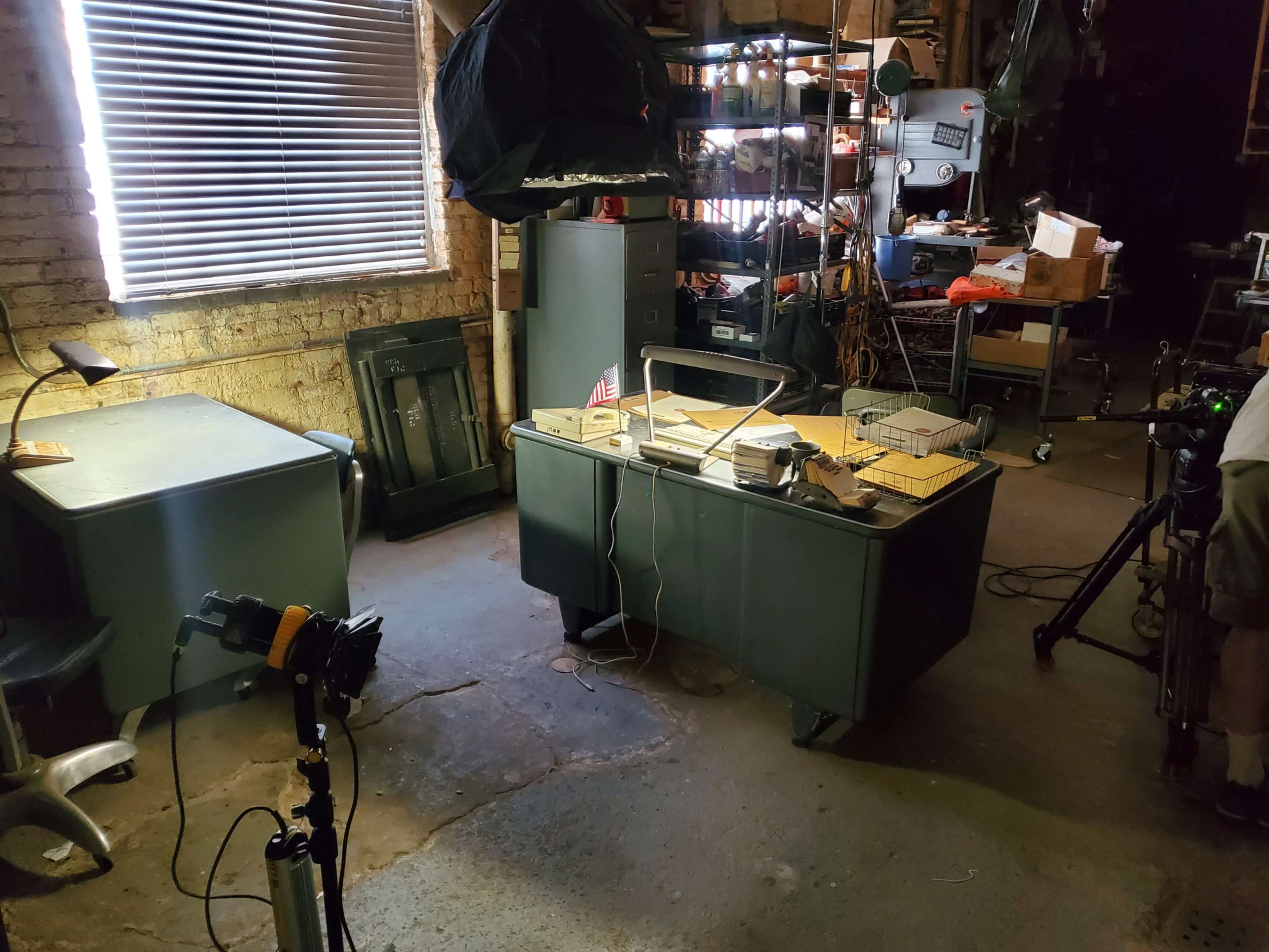 The image shows a cluttered garage or workshop with a metal desk, a few chairs, shelves filled with various items, and a window with blinds partially closed.
