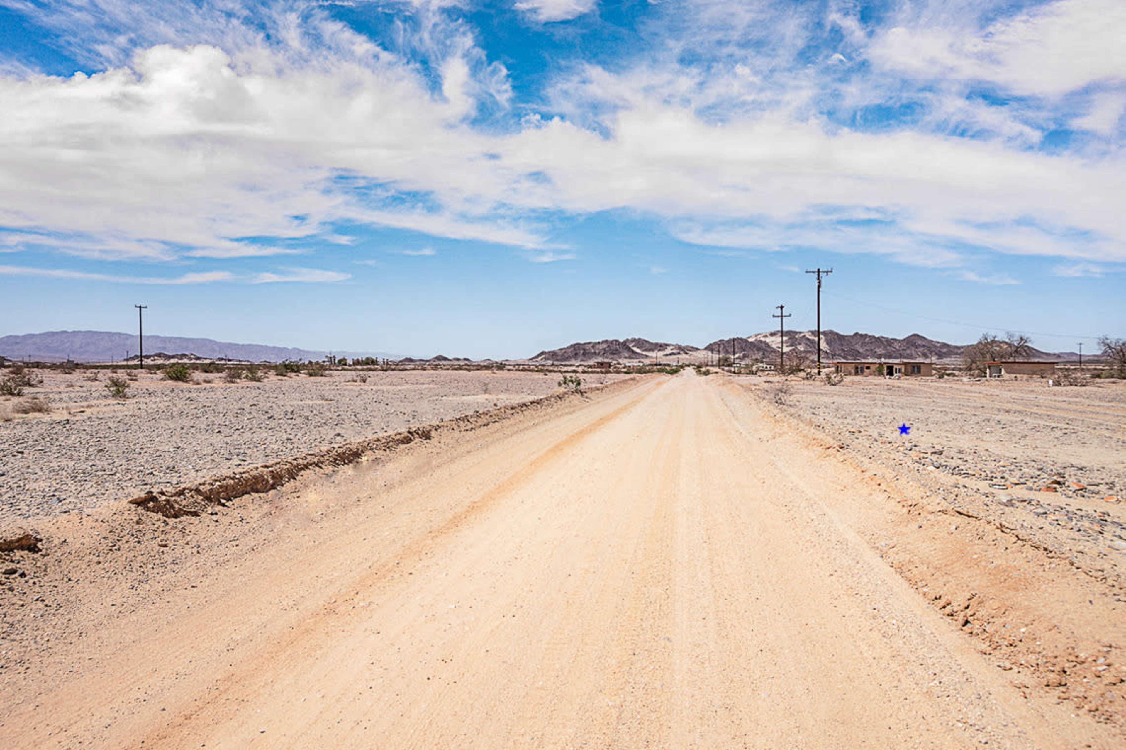 A dirt road stretches through a barren landscape, flanked by sparse vegetation and distant mountains under a blue sky.