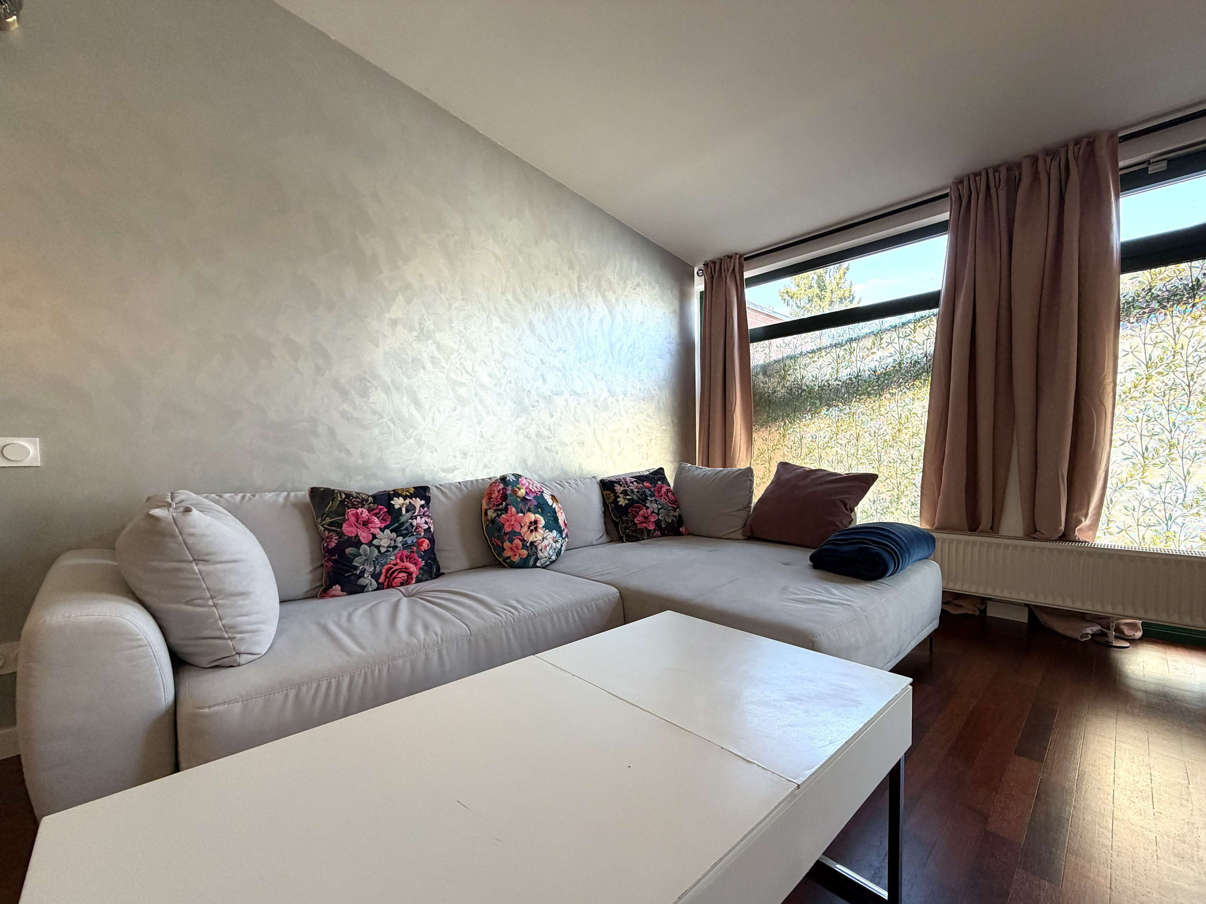 A brightly lit living room features a white sectional sofa adorned with floral decorative pillows and a small white coffee table.