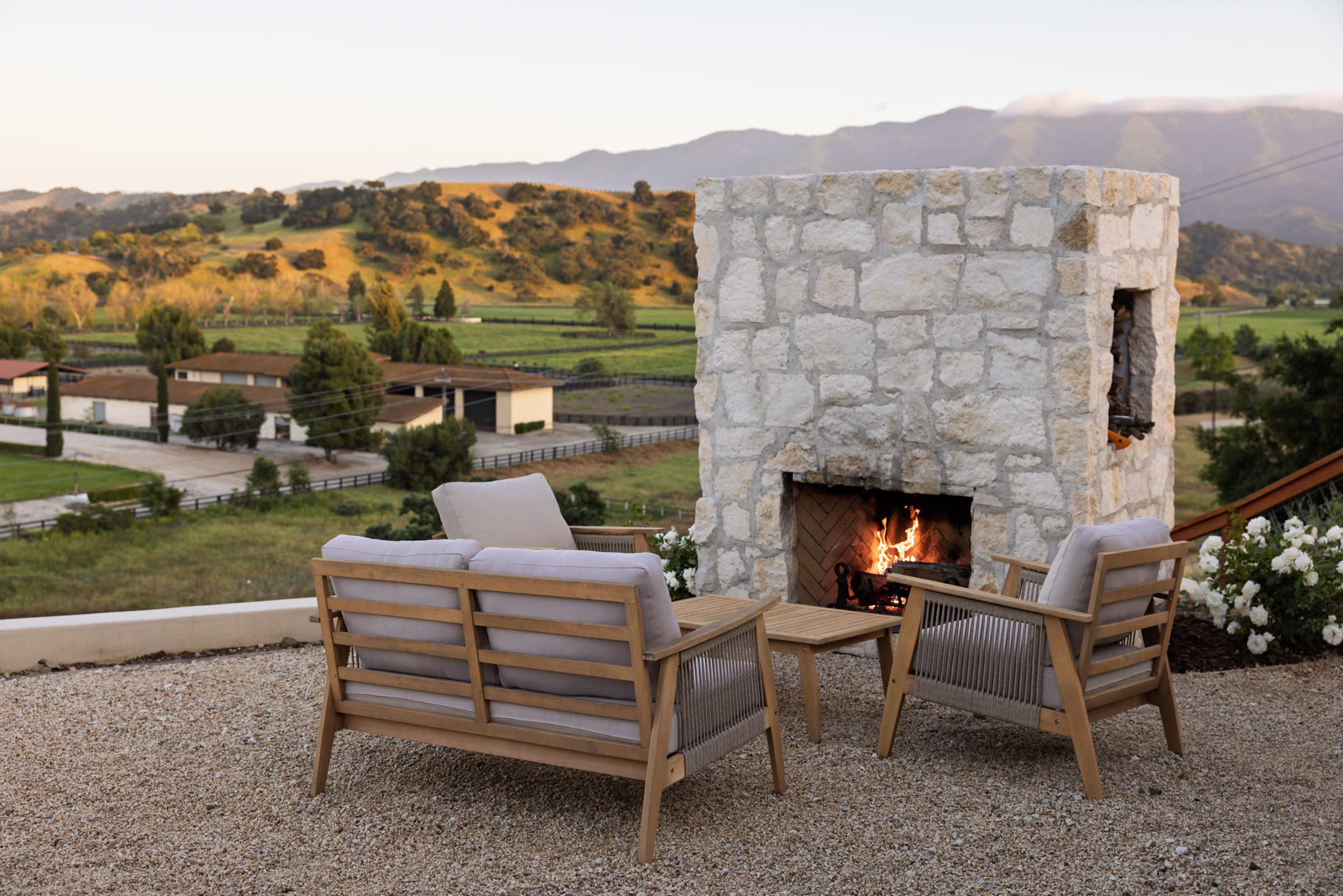 An outdoor seating area with two chairs and a wooden table faces a stone fireplace, surrounded by a scenic landscape of hills and farm buildings.