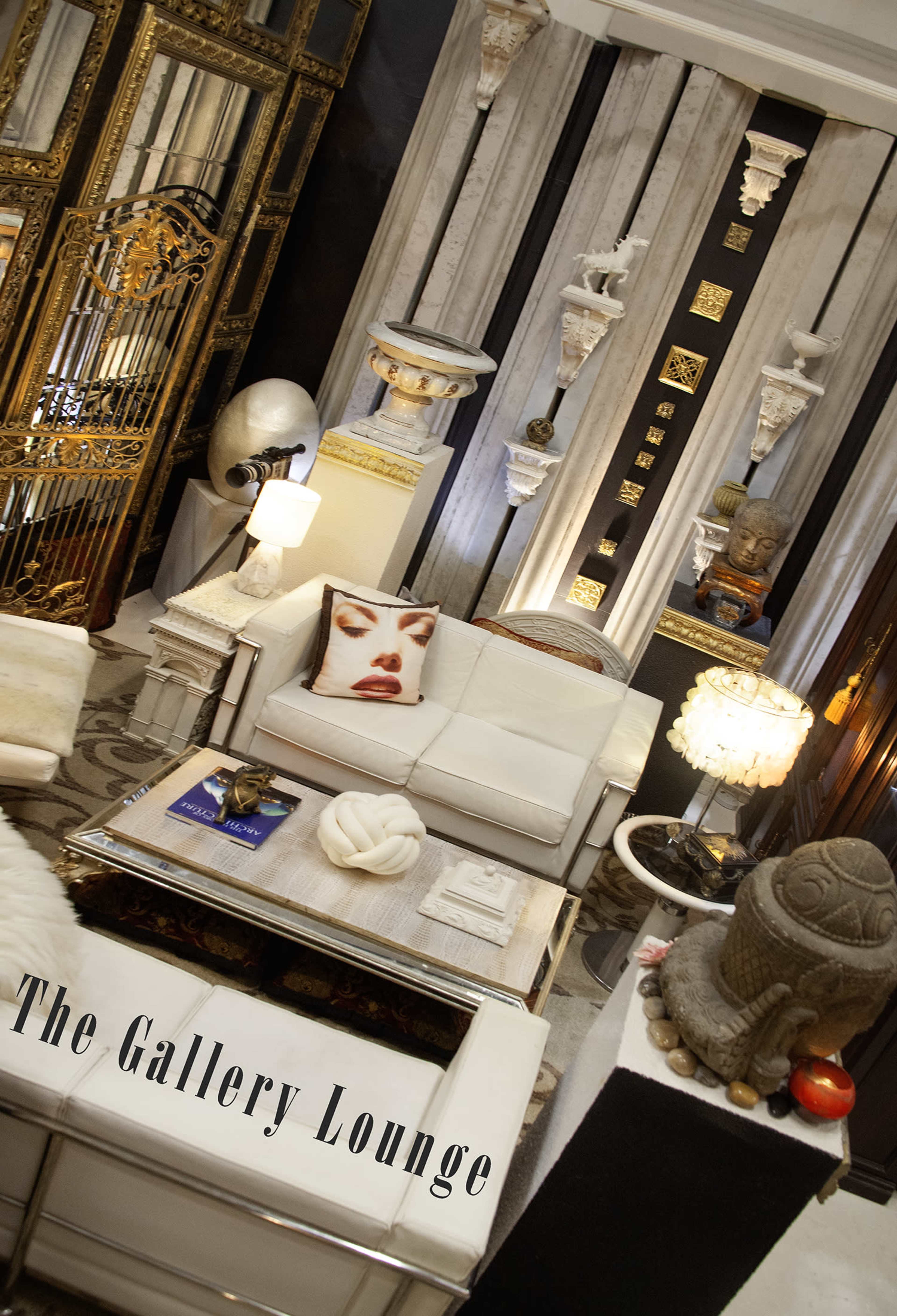 The image shows a stylish lounge area featuring white furniture, decorative sculptures on the walls, and a large pillow with a prominent face design.