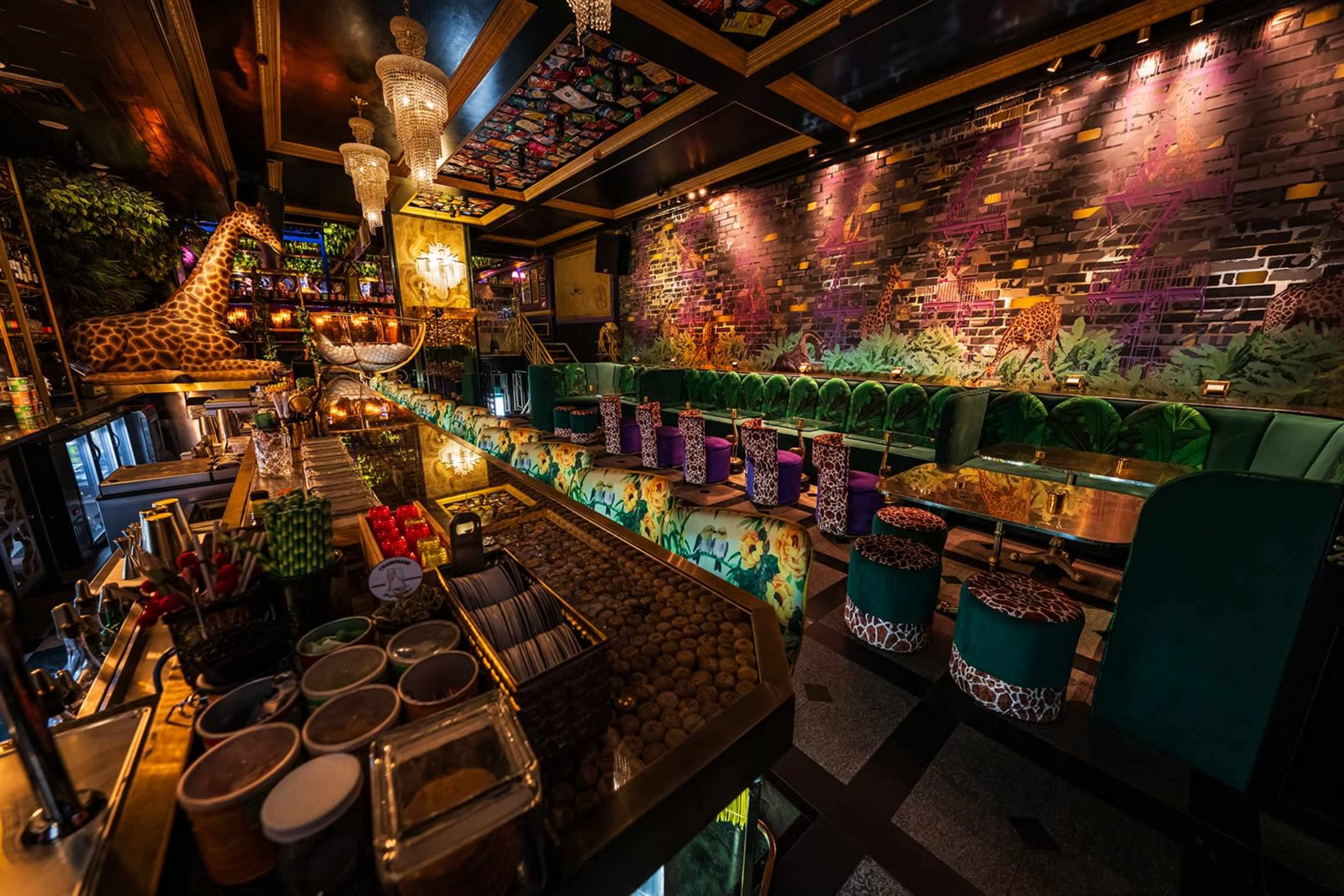The image shows a vibrant restaurant interior featuring a bar, green seating, and colorful wall murals of animals and foliage.