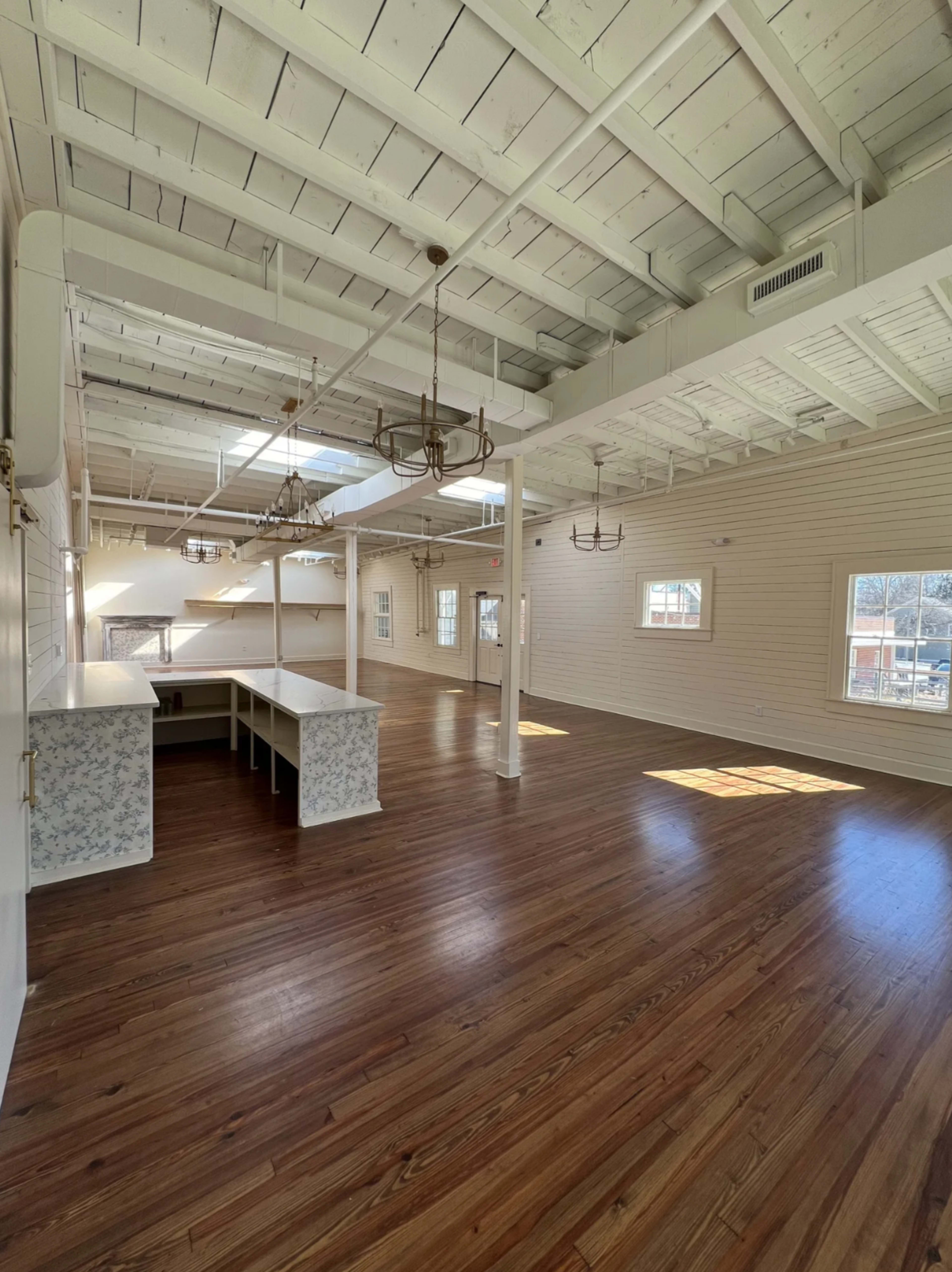The image depicts a spacious, airy room with hardwood flooring, large windows, and a white ceiling with exposed beams.