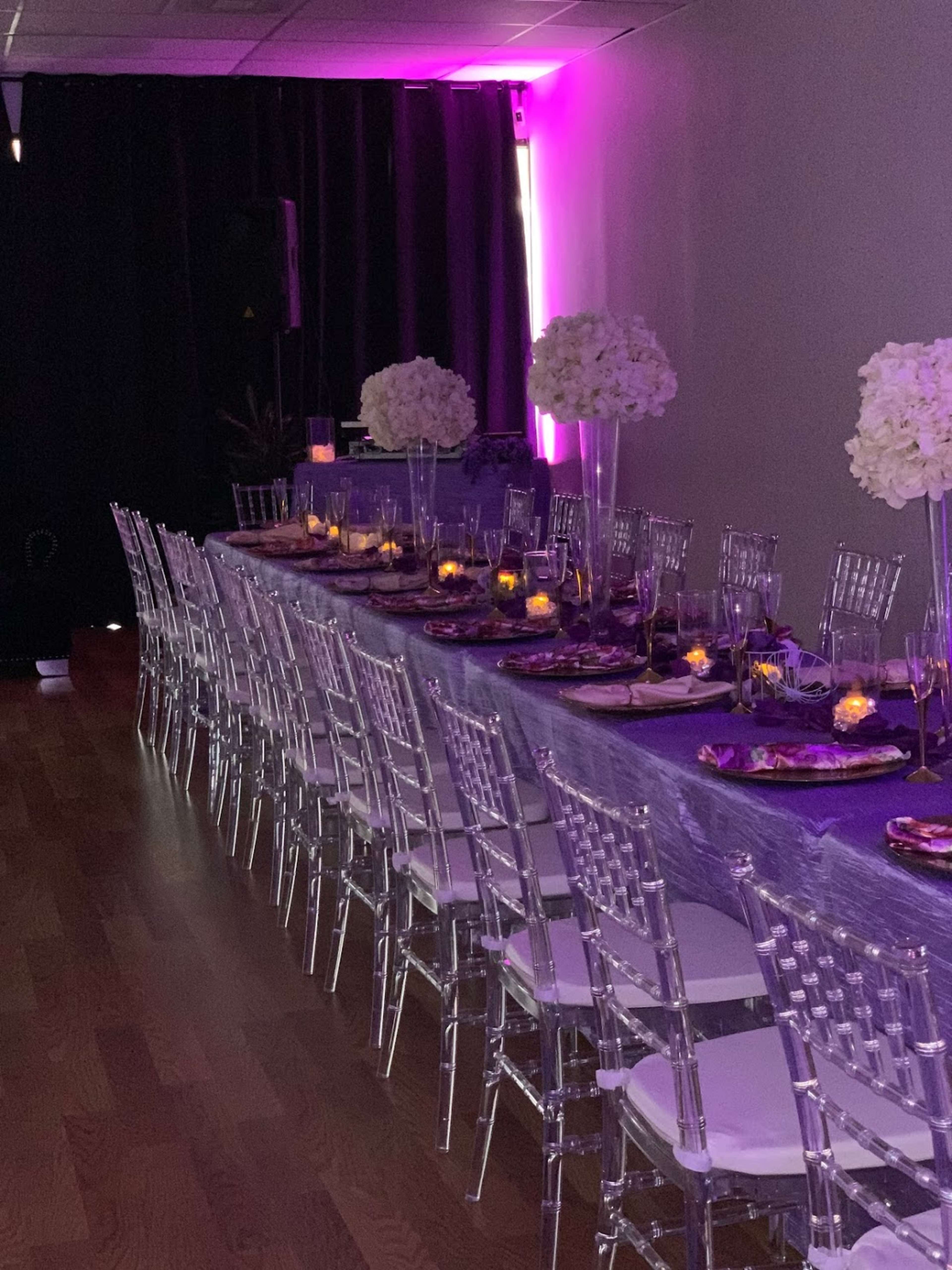 A long dining table is elegantly arranged with transparent chairs and centered floral arrangements, set against a backdrop of purple lighting.