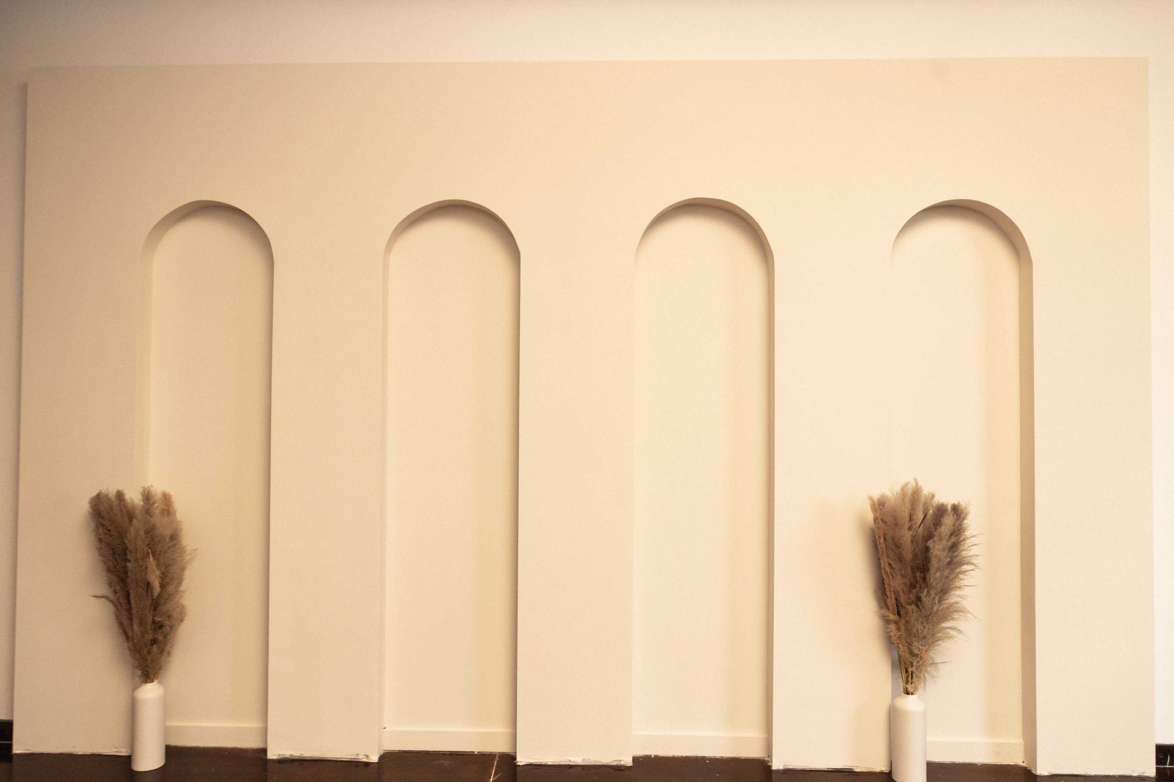 A minimalist wall with three arched niches and two decorative pampas grass arrangements in white vases on either side.