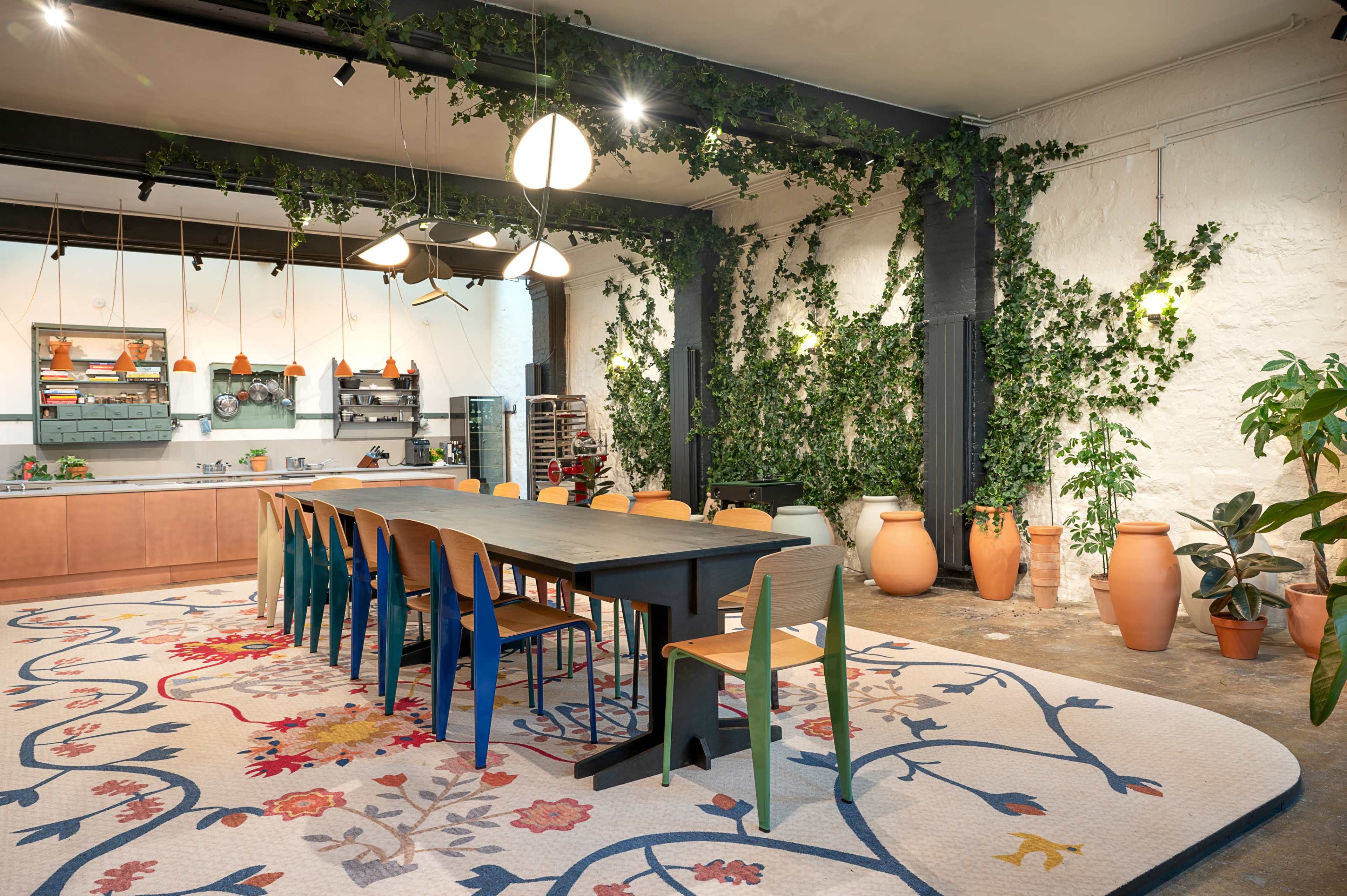 The image shows a modern kitchen and dining area featuring a long table surrounded by colorful chairs, with green plants and decorative pottery integrated into the decor.