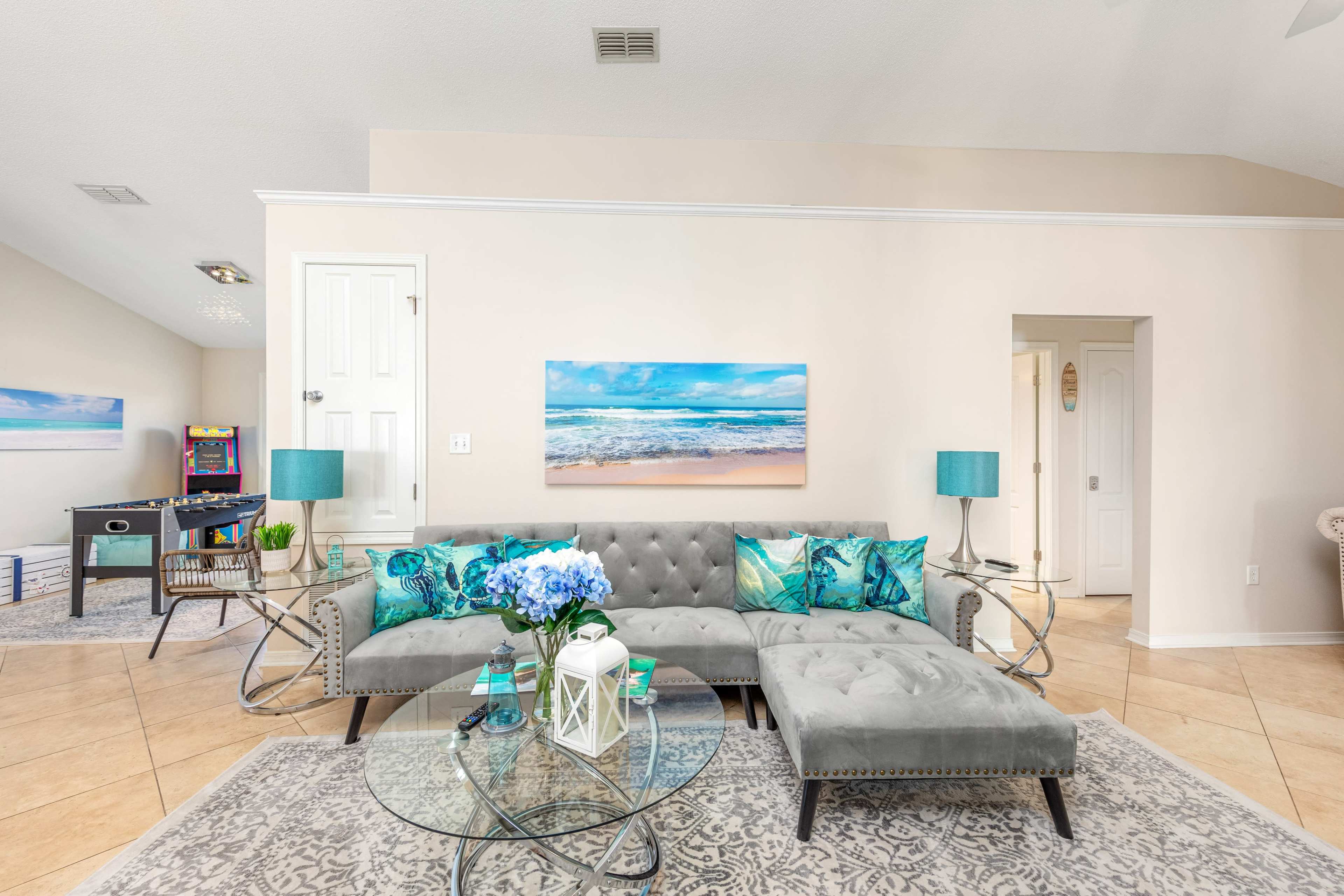 The living room features a tufted gray sofa with blue and green pillows, a glass coffee table with a white decorative lantern, and a large beach-themed painting on the wall.