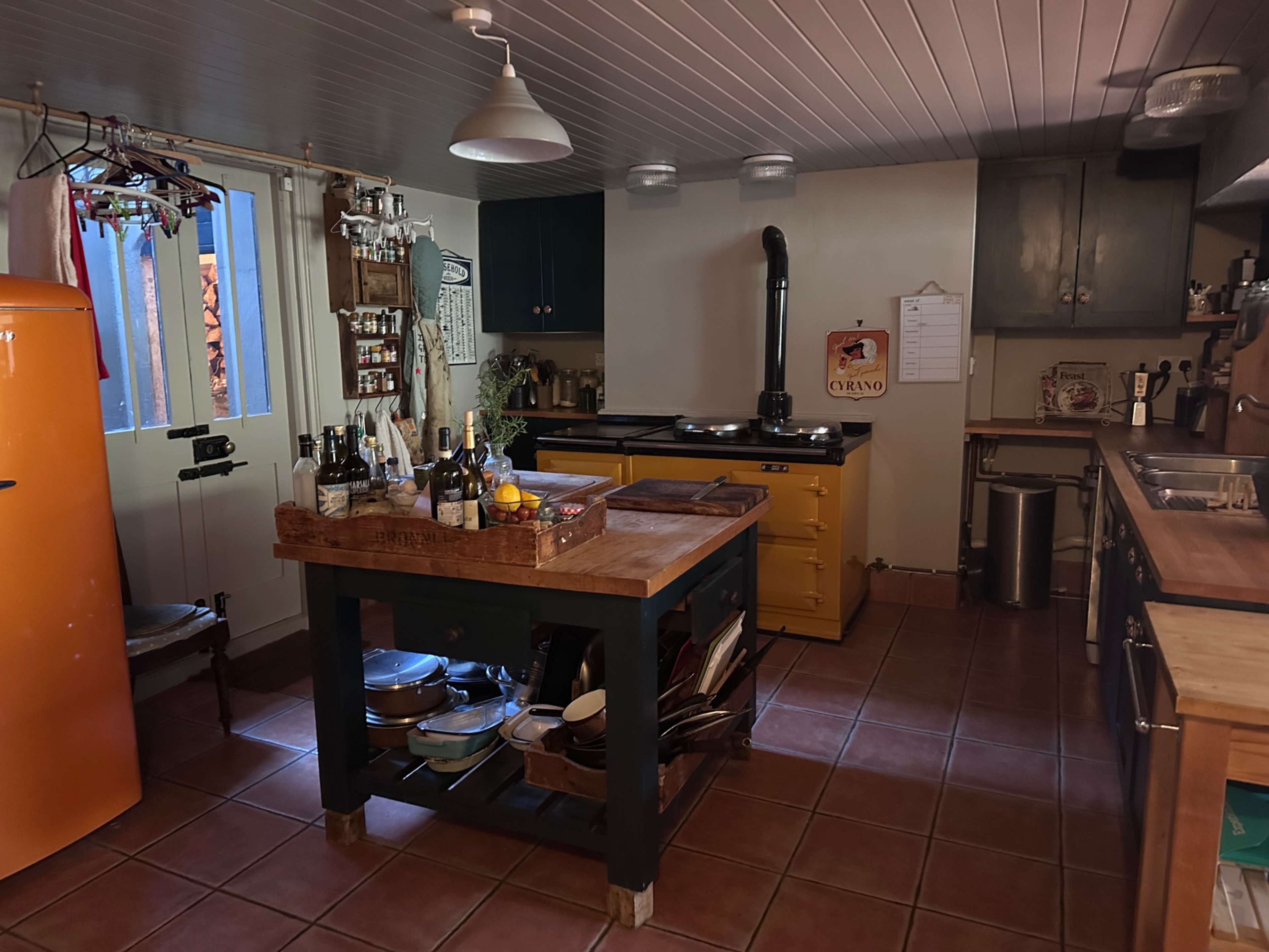 The kitchen features a vibrant orange refrigerator, a wooden island with a basket of fruit, and blue cabinets alongside a yellow stove.