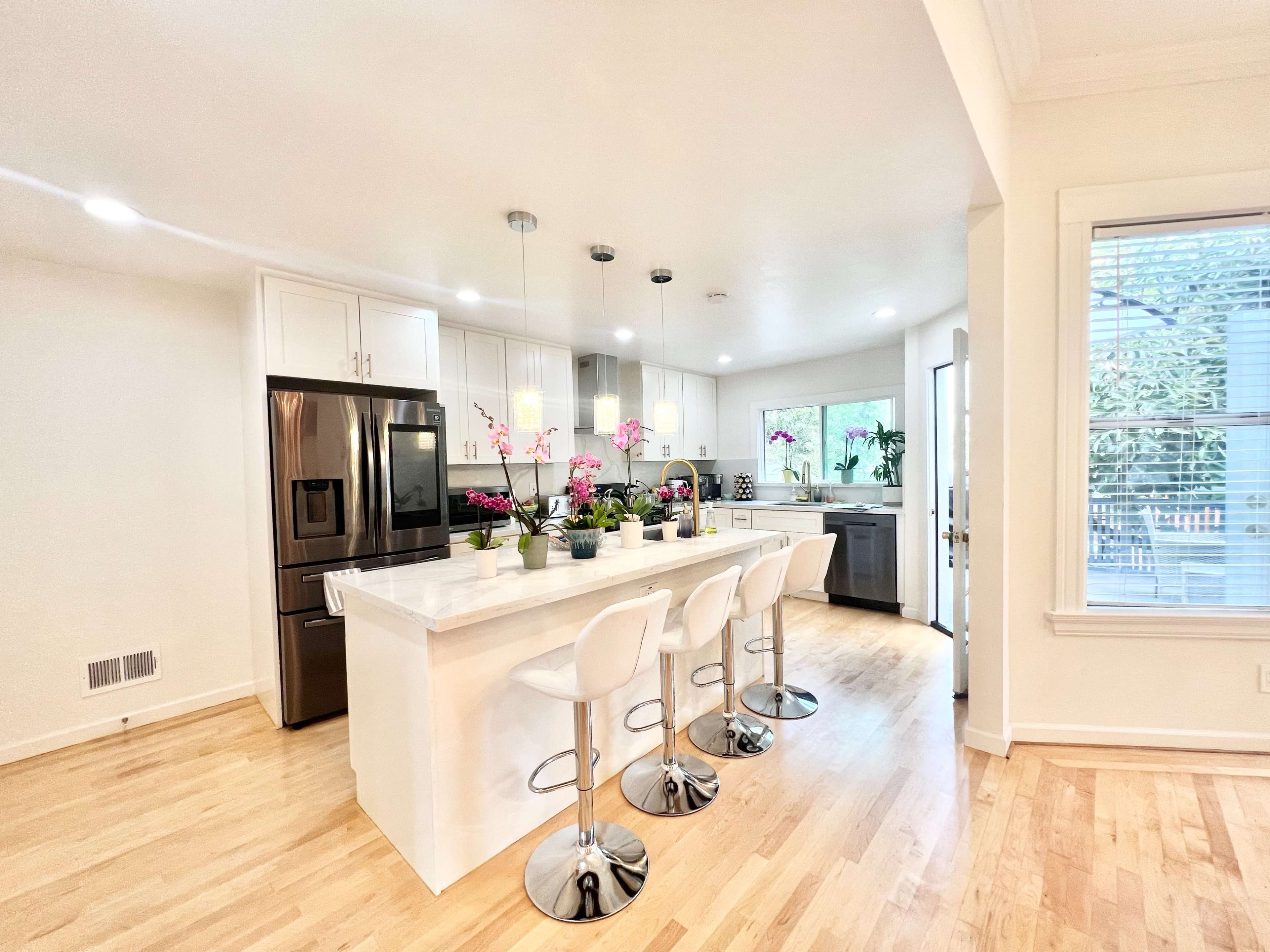 A modern kitchen features white cabinetry, a marble island with bar stools, and natural light coming through large windows.