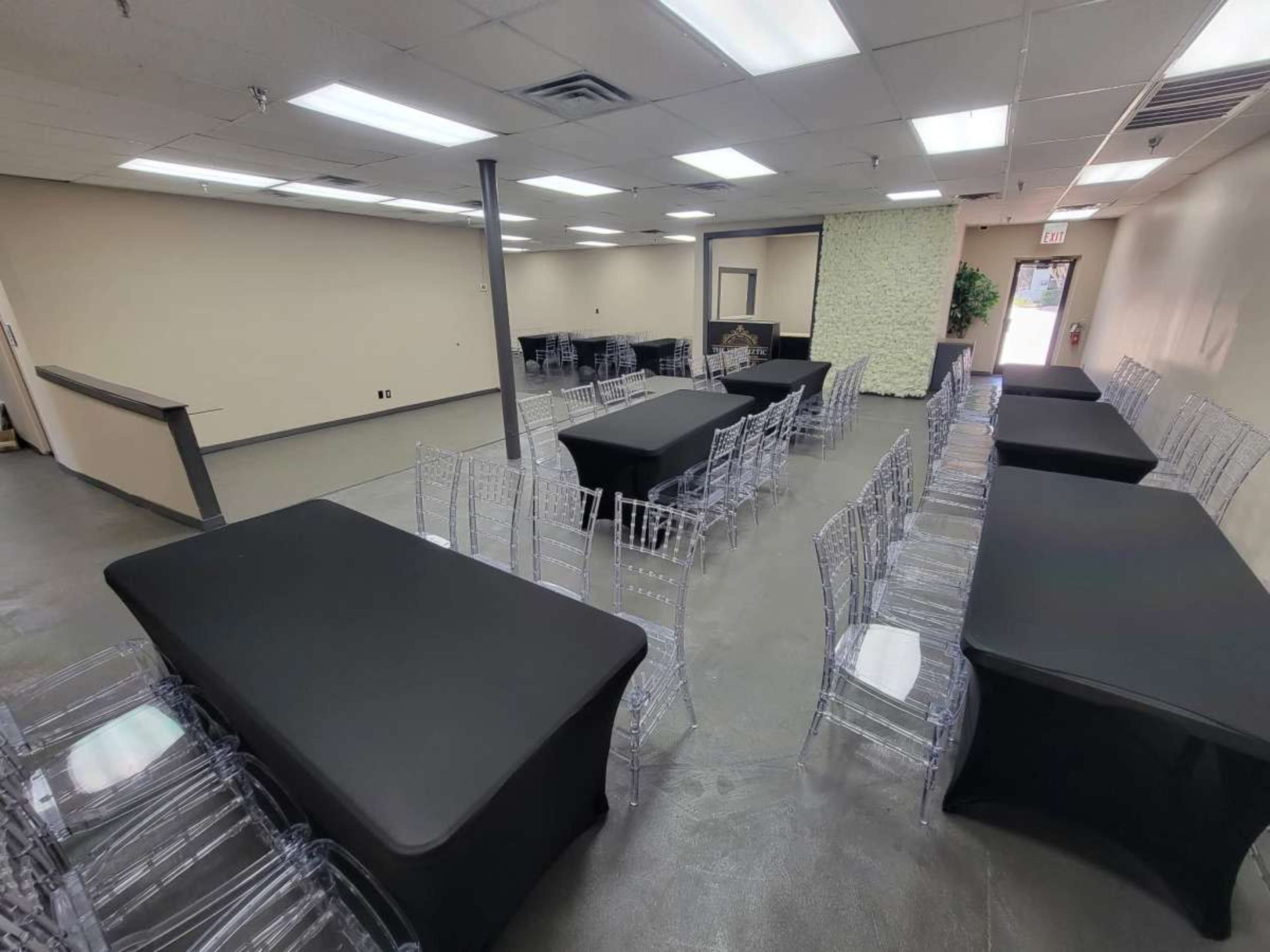 The image shows a spacious room arranged with multiple tables covered in black tablecloths and clear acrylic chairs set in a grid pattern.